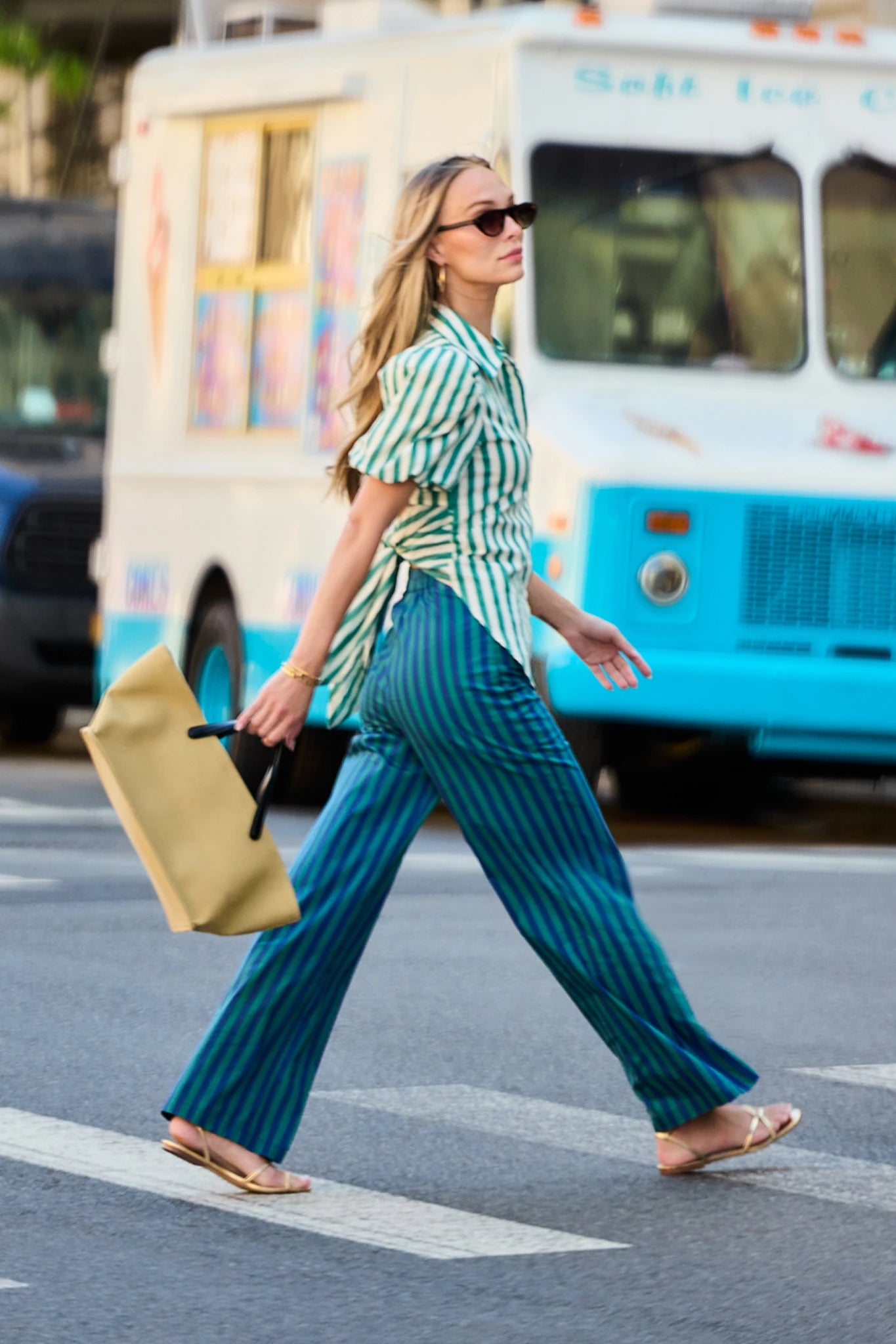 A woman walks across a street crosswalk wearing a green and white striped short-sleeve shirt and teal blue pinstriped wide-leg pants, carrying a tan bag. A white and blue ice cream truck is blurred in the background.