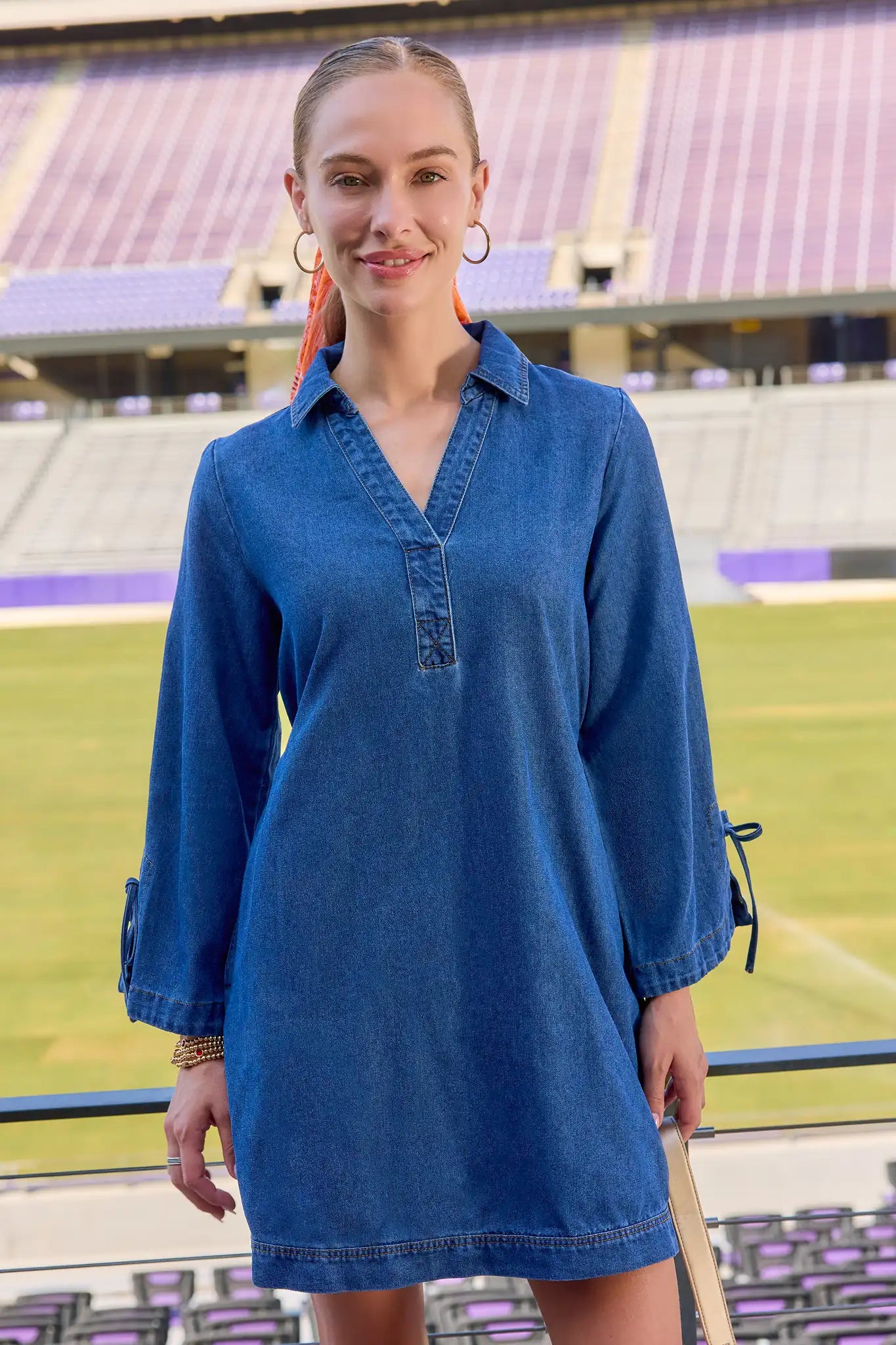 A woman wearing a blue denim long-sleeve collared shirt dress stands in front of a stadium background.