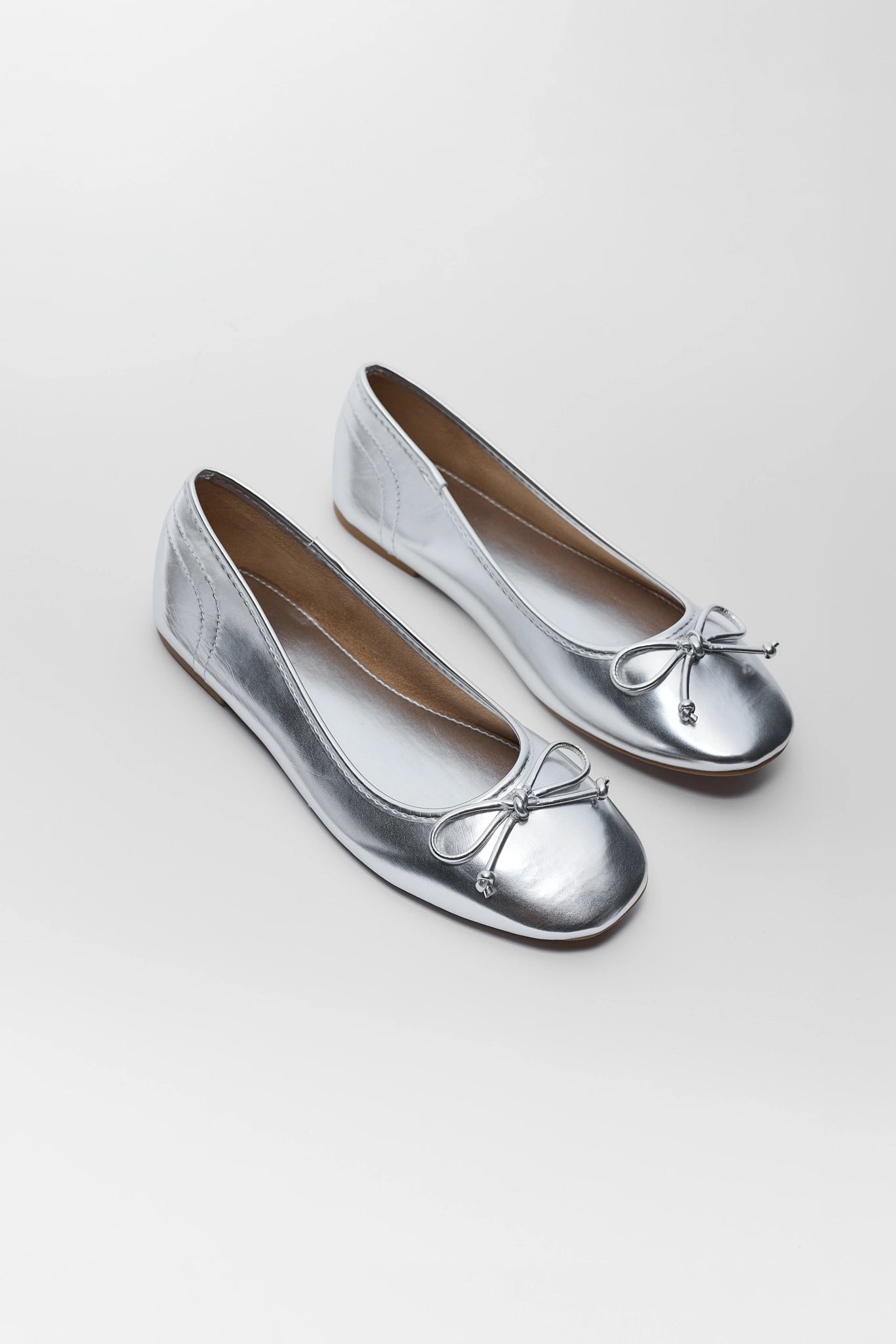 A pair of metallic silver ballet flats with small silver bow details on the toes, shown on a light background.