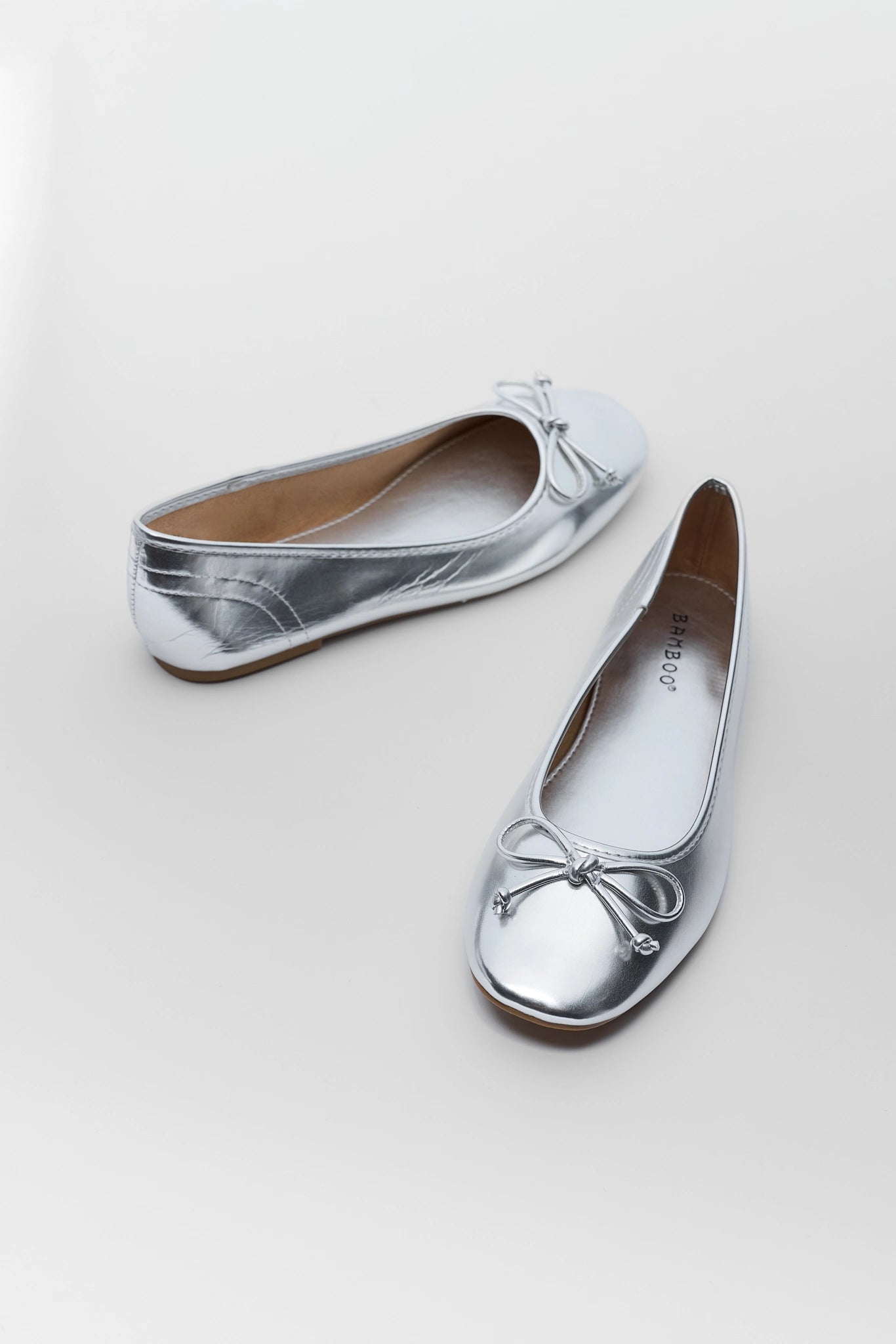 A pair of metallic silver ballet flats with small bow details on the toes, displayed on a light gray background.