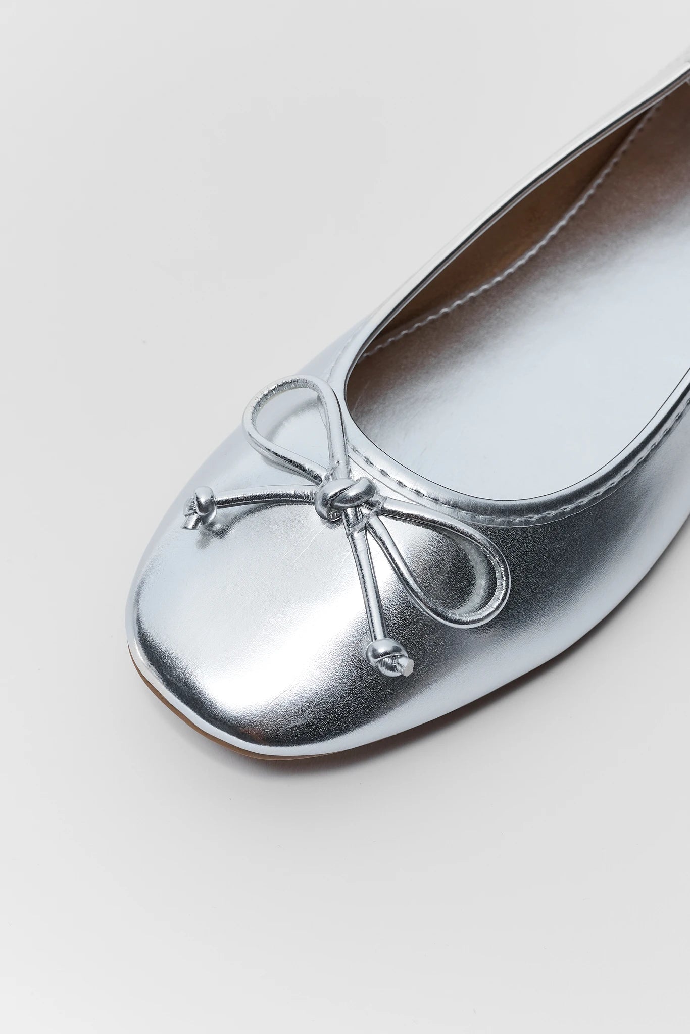 A close-up of a metallic silver ballet flat shoe with a decorative silver bow on the toe, set against a clean light gray background.