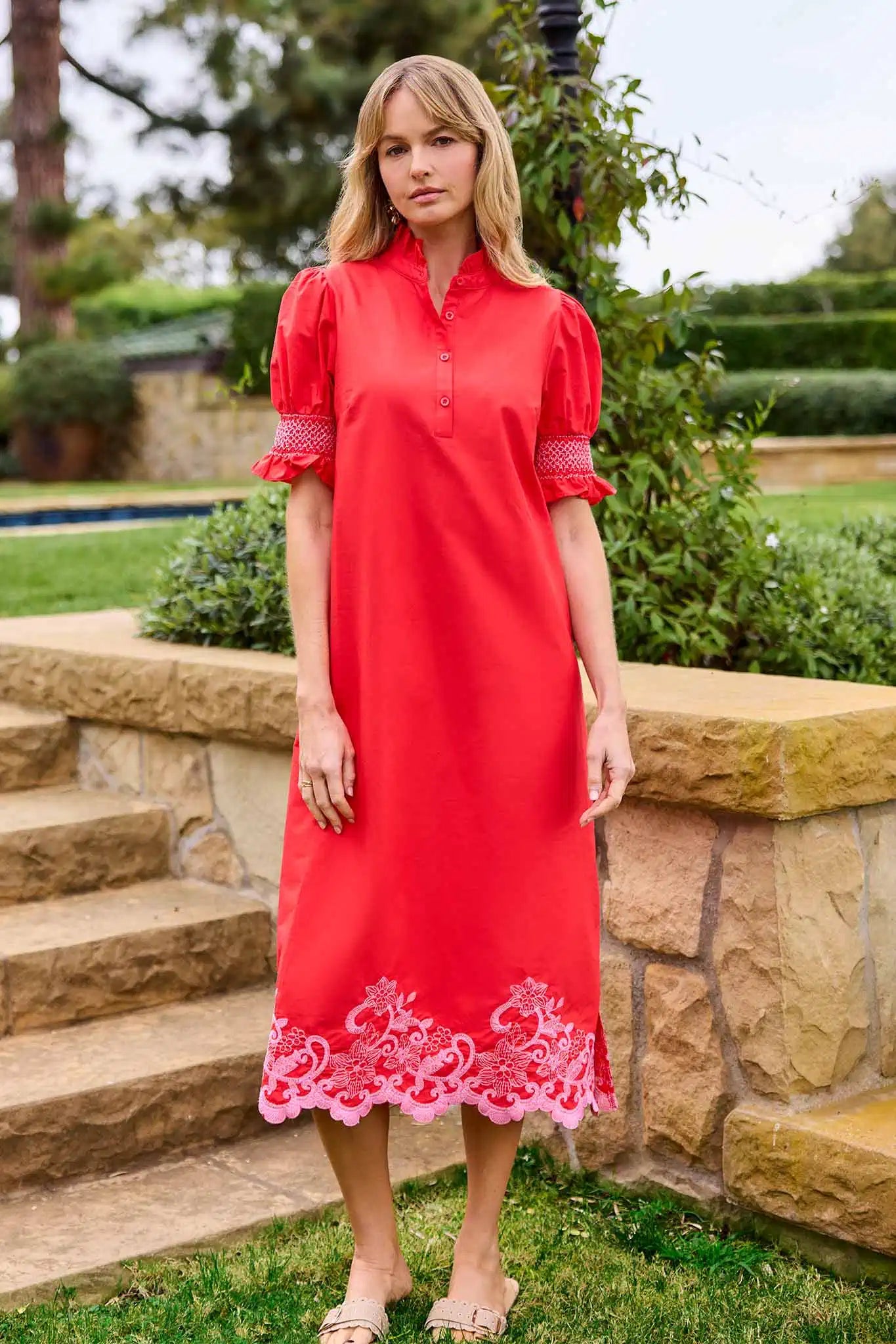 Woman wearing a red dress with lace details standing outdoors near stone steps and greenery.