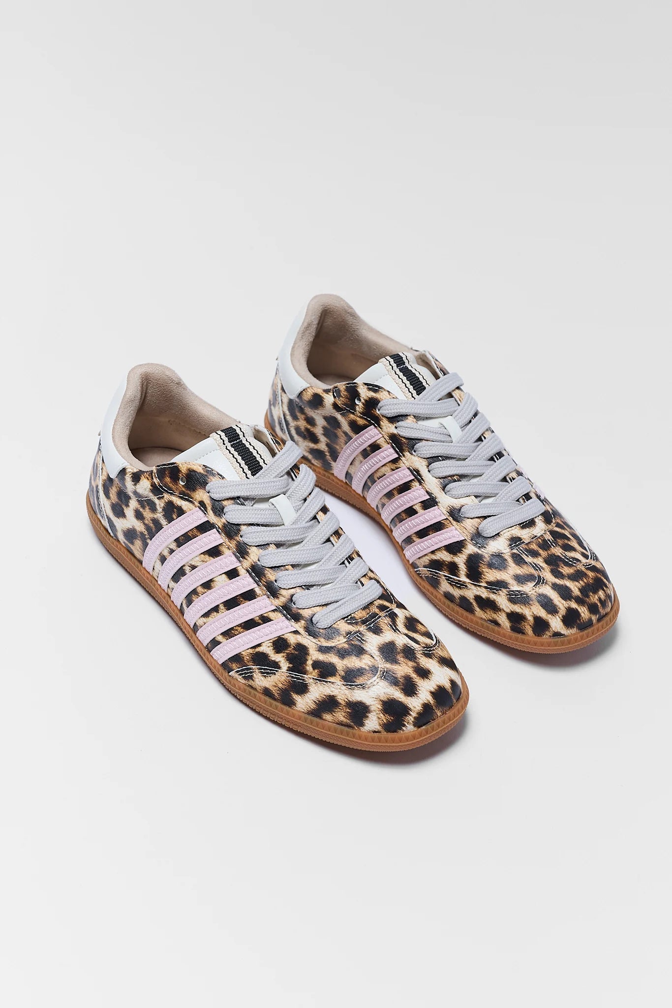 A pair of low-top sneakers featuring a leopard print pattern on a tan base, with pale pink laces and white eyelets, photographed on a light gray background.