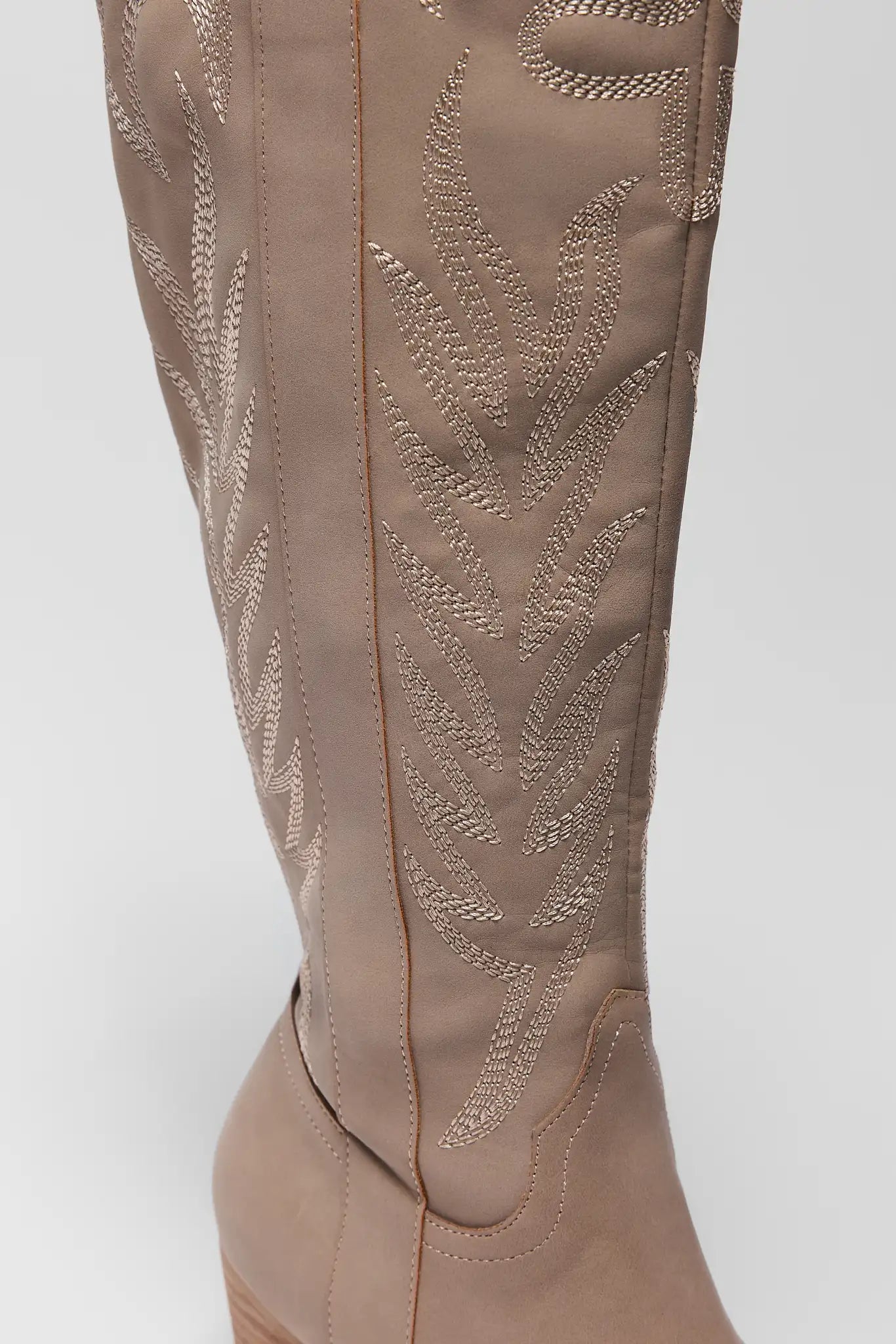 Close-up of a tall western-style boot in taupe suede with metallic embroidered leaf patterns on the shaft, shown against a light gray background.