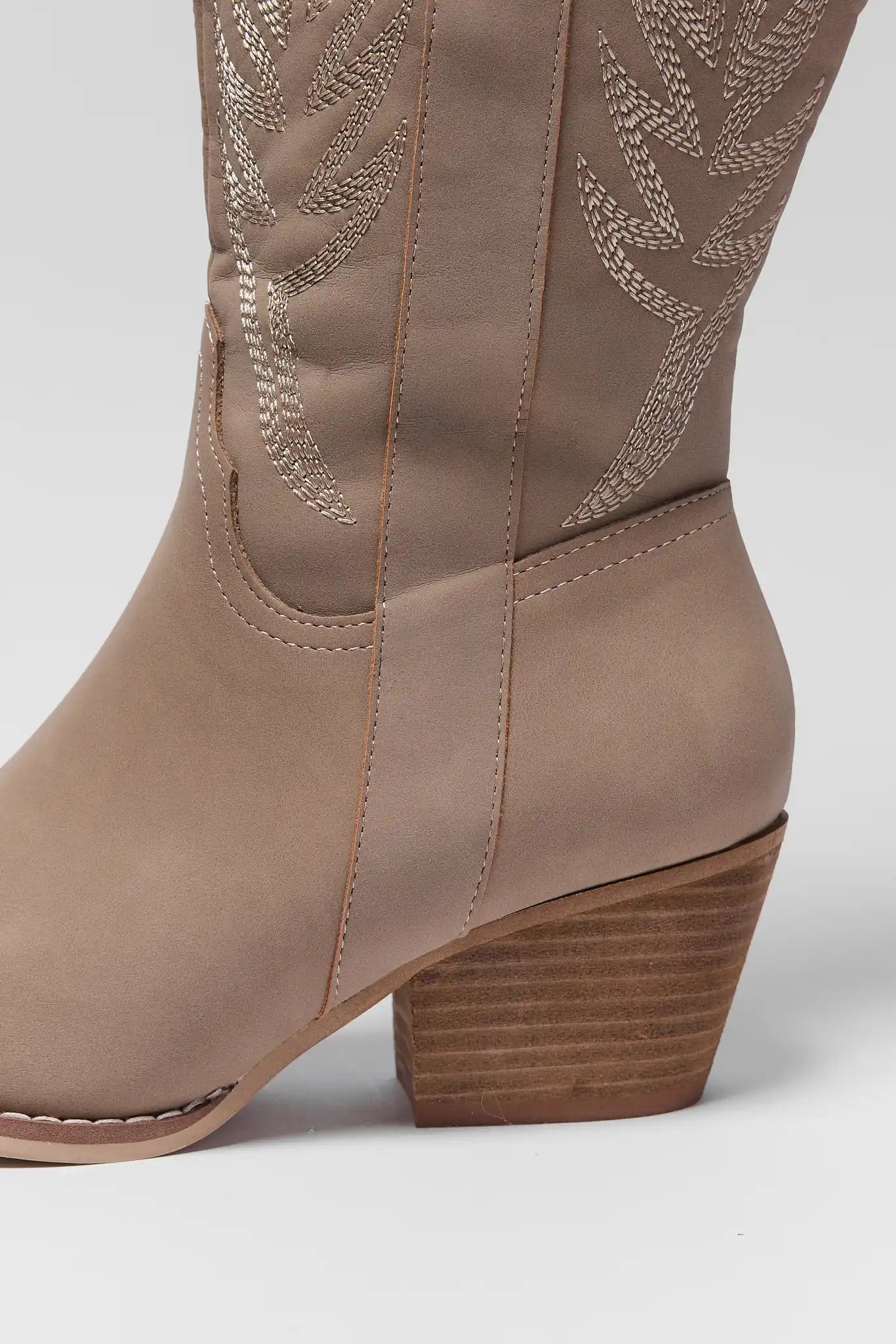 A close-up side view of a taupe western-style boot with decorative metallic embroidery on the shaft and a stacked brown block heel, set against a plain light gray background.