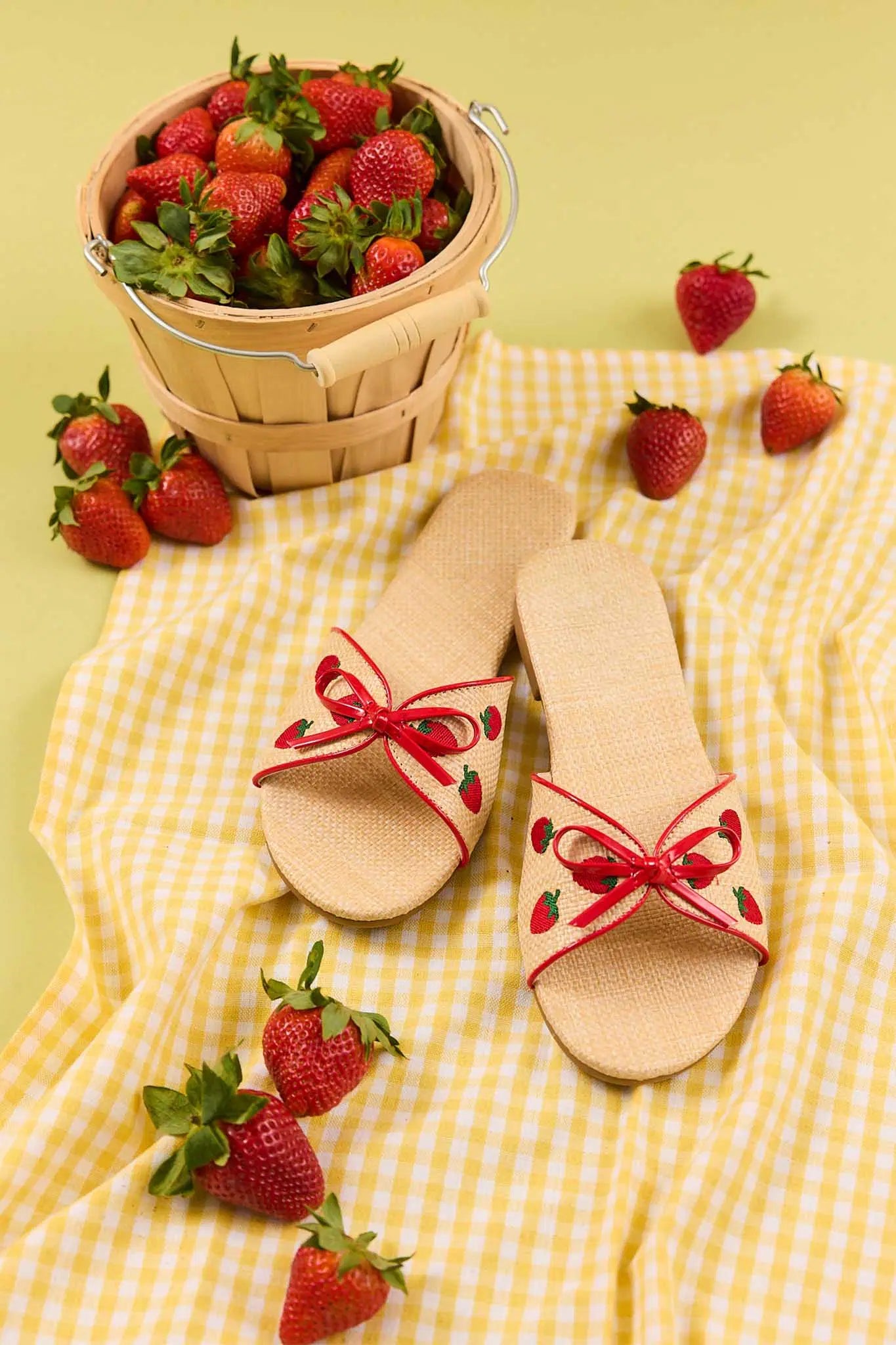 Beige sandals with red strawberry details on a yellow checkered cloth with strawberries around.