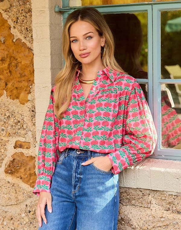 Woman wearing a pink floral shirt and blue jeans standing in front of a stone wall.
