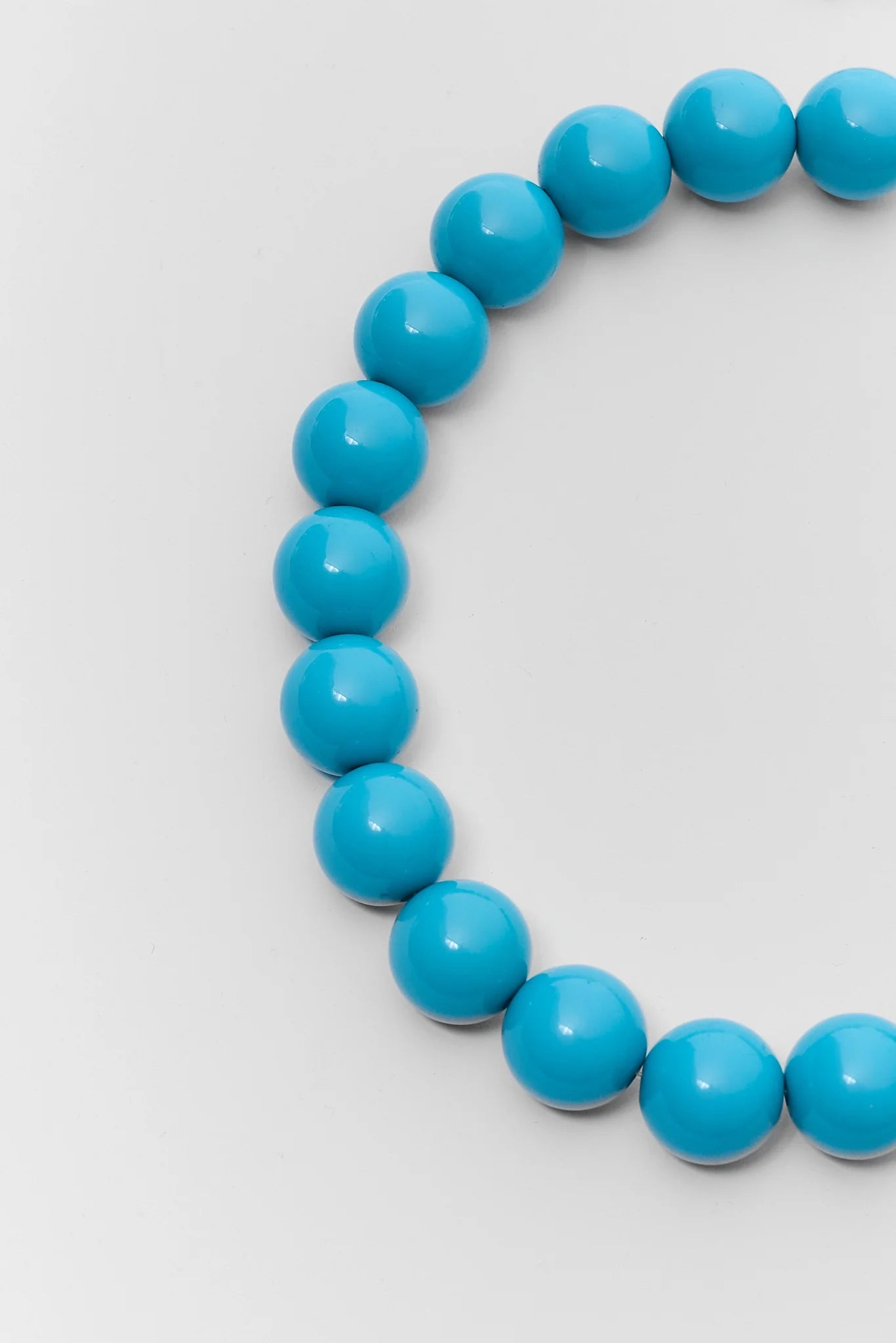 A close-up of a beaded necklace made of glossy, round turquoise-blue beads, arranged in a curve against a light gray background.