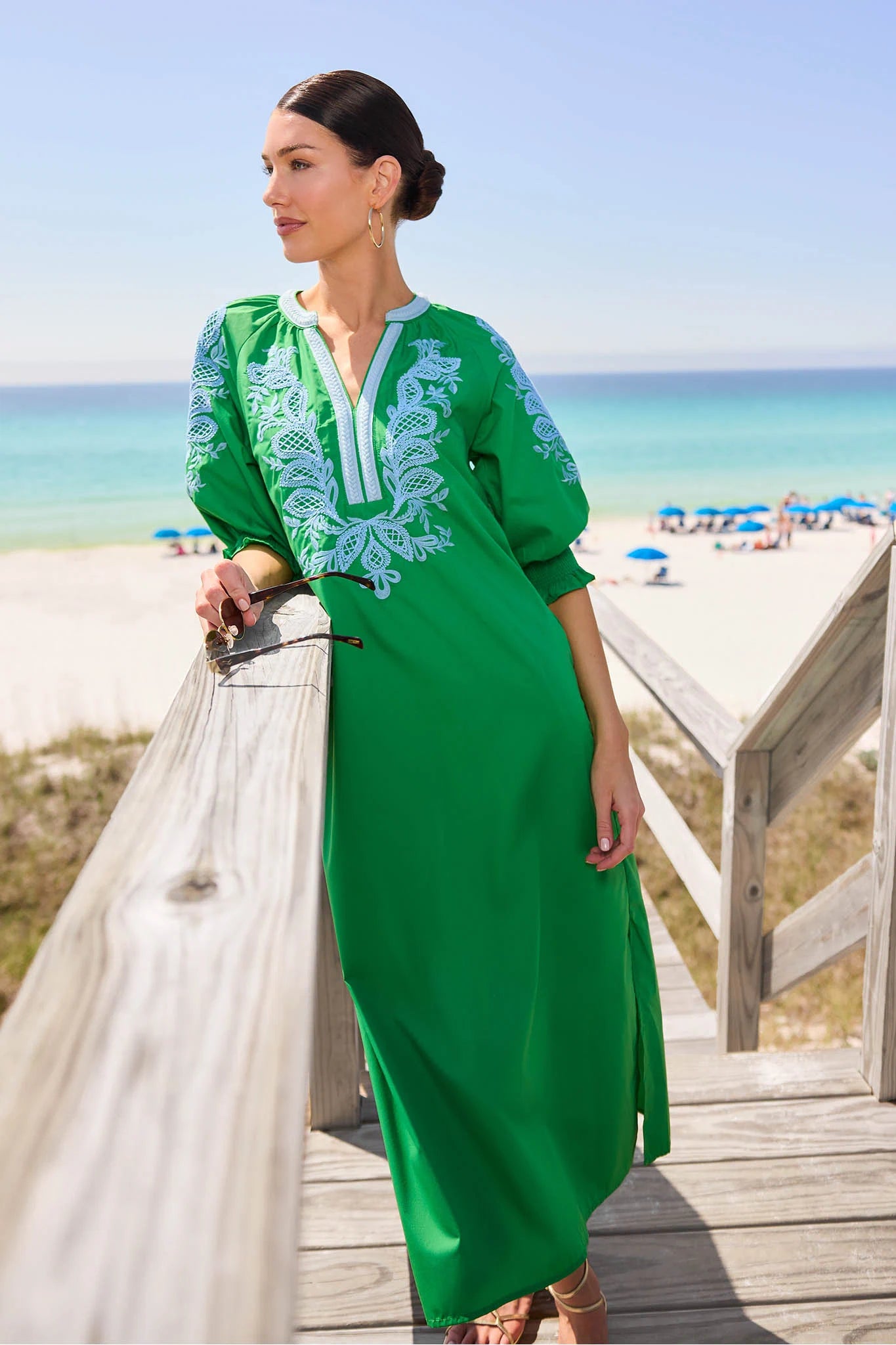 Woman in a green dress with white embroidery standing on a beach.