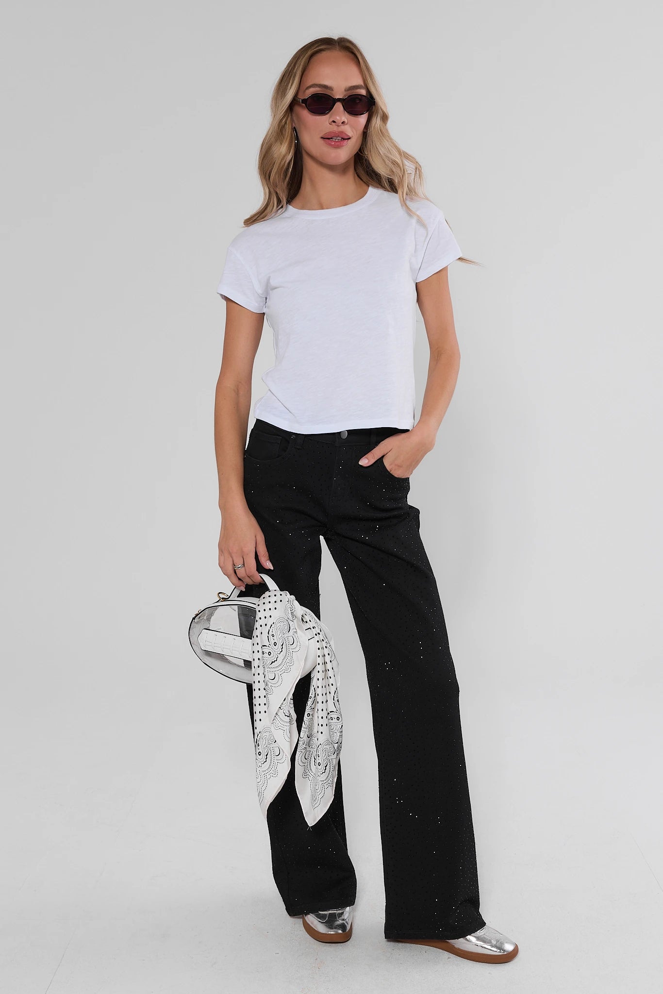 A woman modeling a white short-sleeve t-shirt, black wide-leg pants, and silver shoes, holding a black and white patterned scarf, against a light gray studio background.