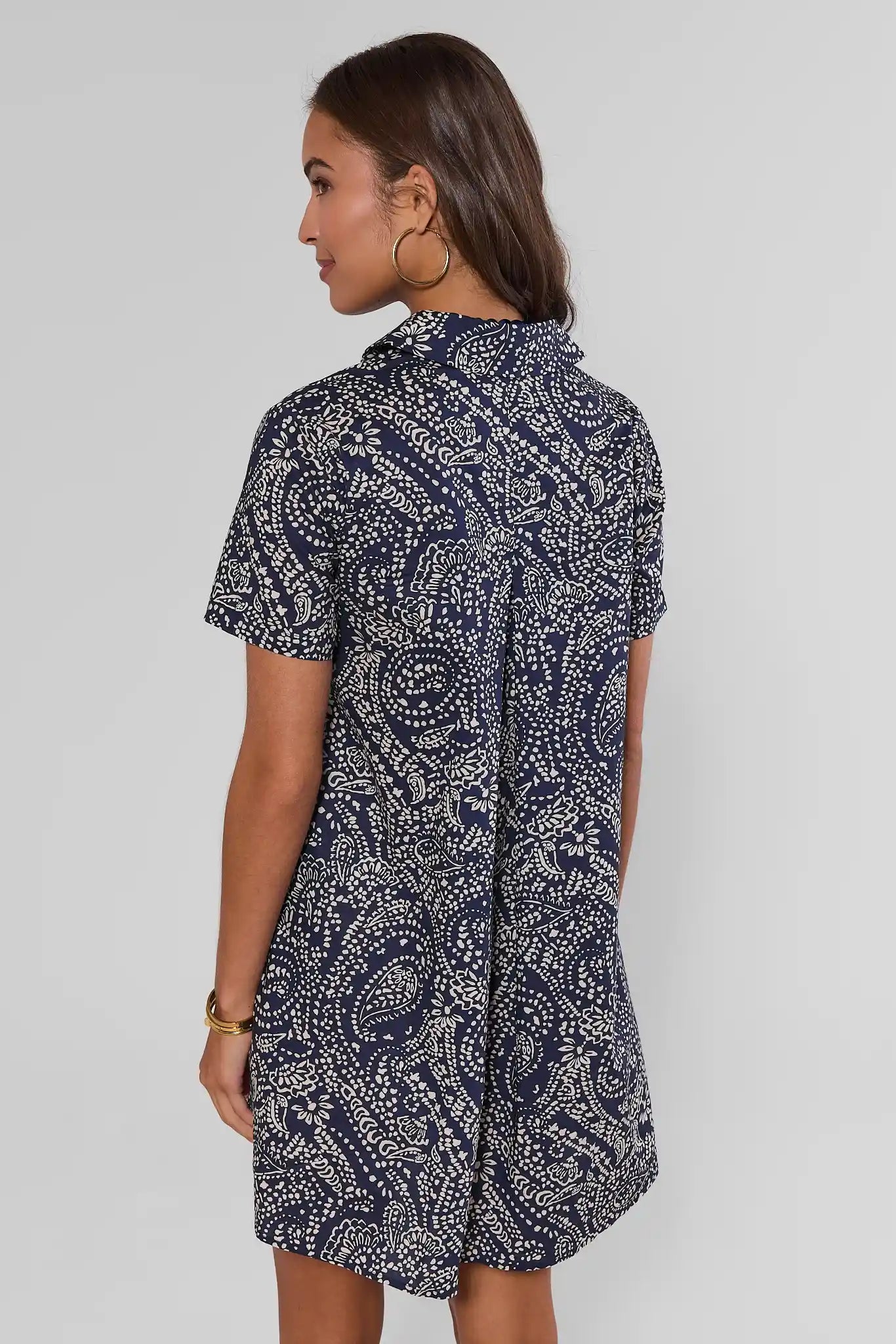A woman shown from behind modeling a short-sleeve mini dress with a navy and white paisley pattern, paired with white sneakers and a dark blue beaded necklace, against a light gray studio background.