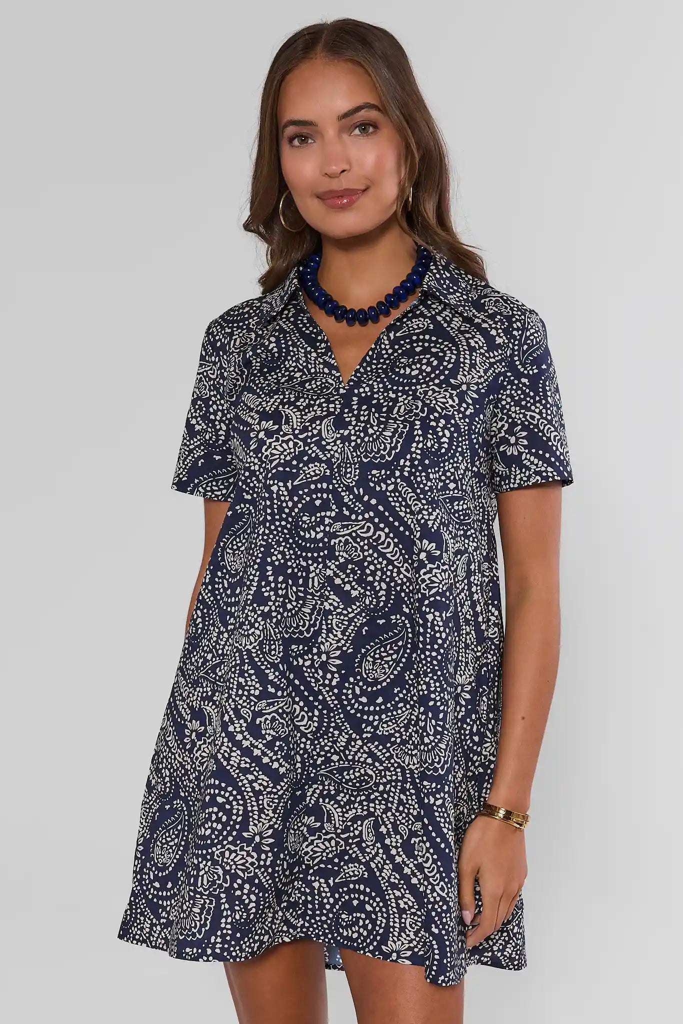 A woman walking modeling a short-sleeve mini dress with a navy and white paisley pattern, paired with white sneakers and a dark blue beaded necklace, against a light gray studio background.