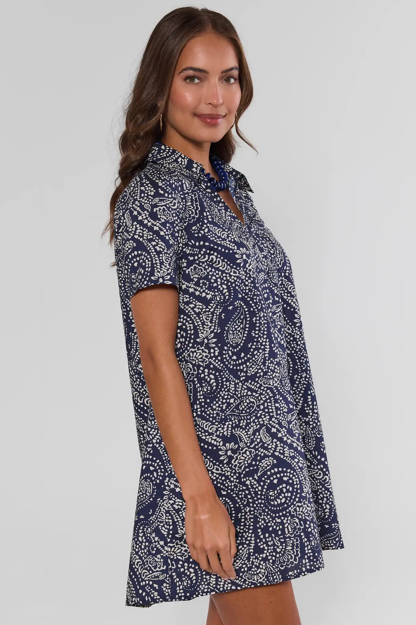 A woman from the side modeling a short-sleeve mini dress with a navy and white paisley pattern, paired with white sneakers and a dark blue beaded necklace, against a light gray studio background.