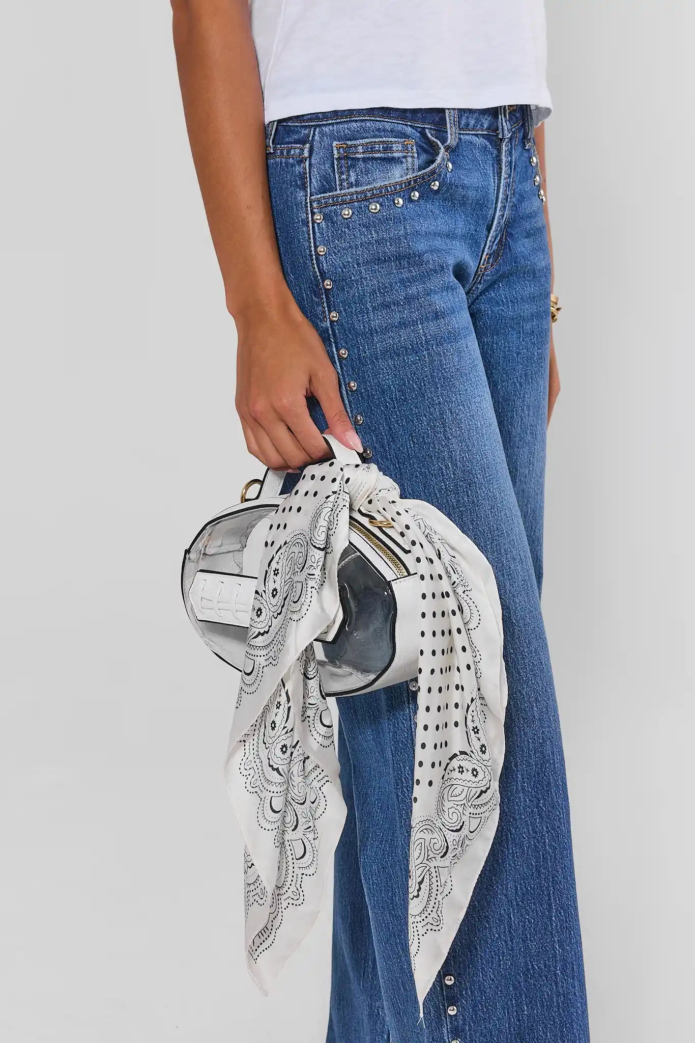 A person wearing blue jeans and a white top holds a small metallic silver handbag with a white patterned scarf tied around it, against a light gray studio background.