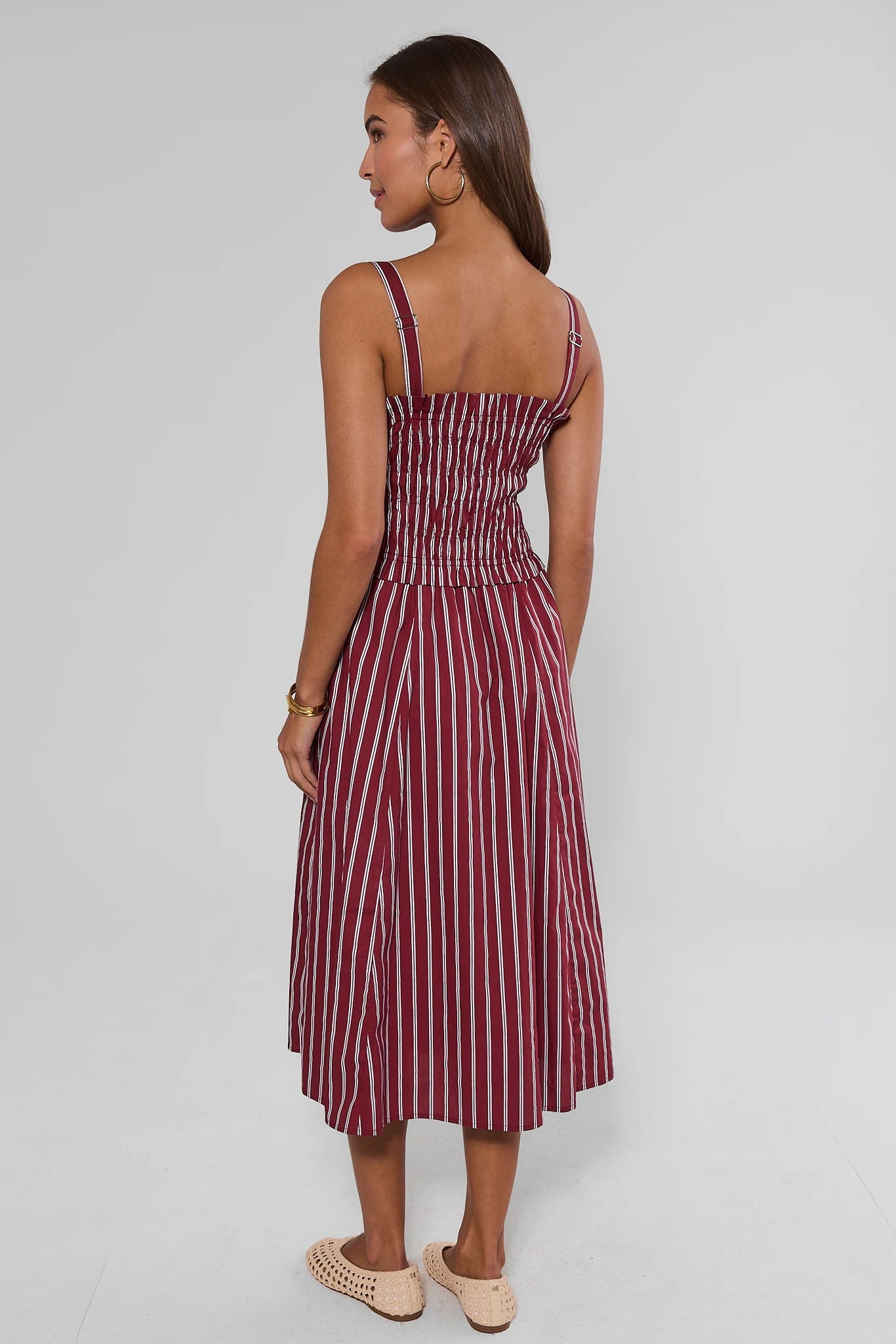 A woman shown from behind modeling a sleeveless midi dress with thin shoulder straps, a smocked bodice, and vertical red and white stripes, set against a plain light gray studio background.