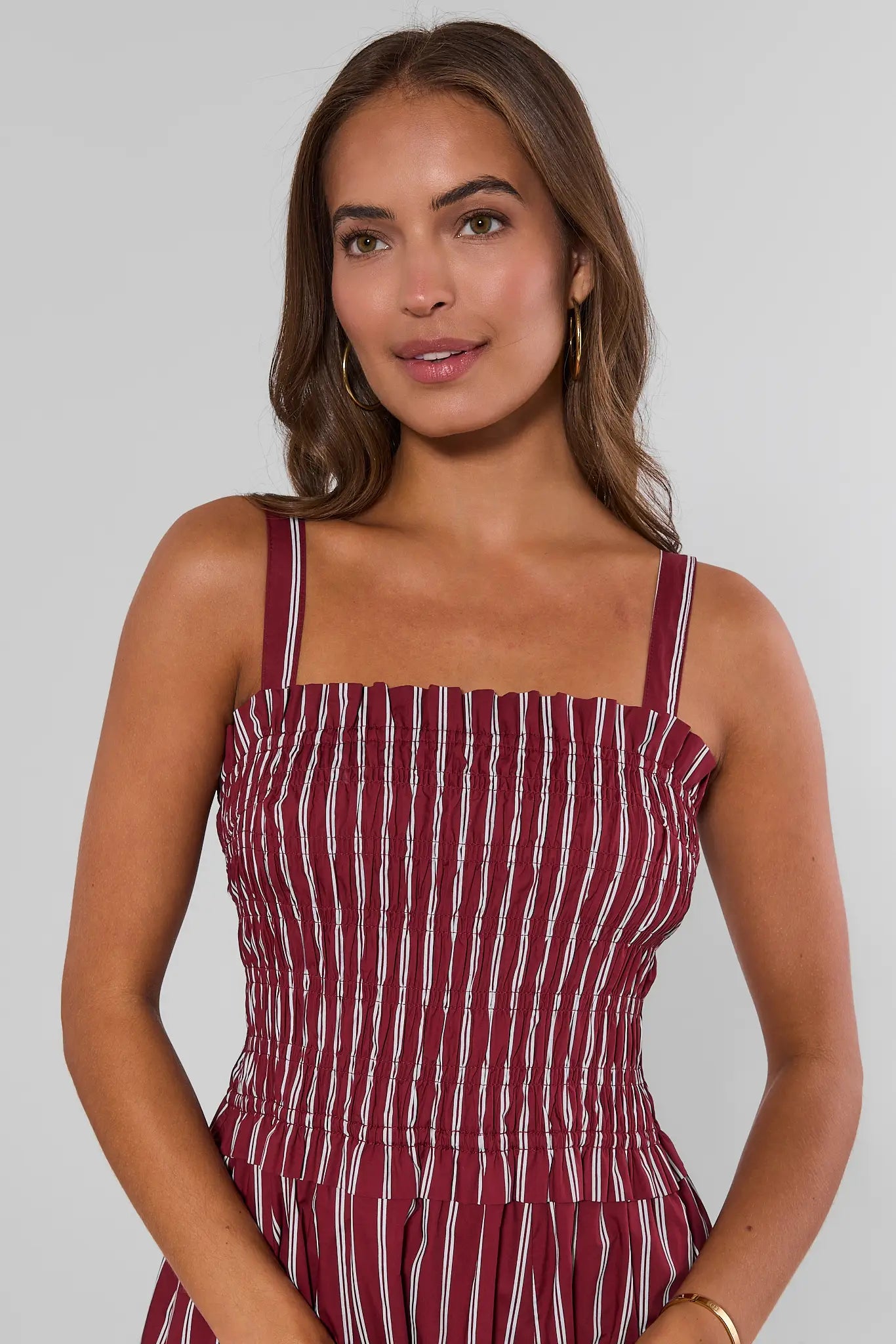 A close up woman modeling a sleeveless midi dress with thin shoulder straps, a smocked bodice, and vertical red and white stripes, set against a plain light gray studio background.