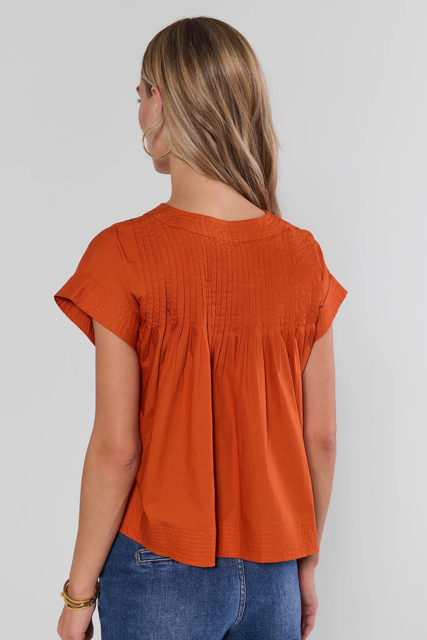 A woman shown from behind wearing a bright orange short-sleeve peplum top with a V-neck and button placket, paired with blue jeans, against a gray studio background.
