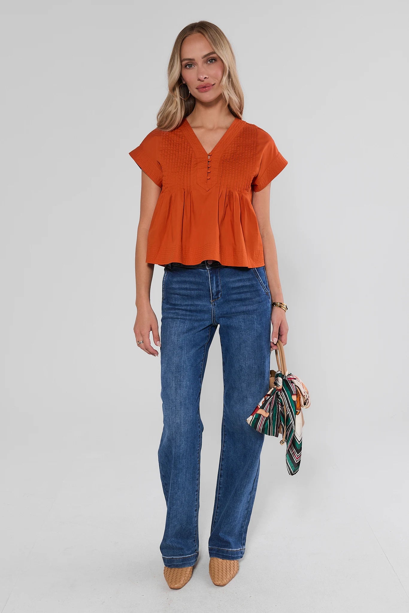 A woman wearing a bright orange short-sleeve peplum top with a V-neck and button placket, paired with jeans, against a light background.