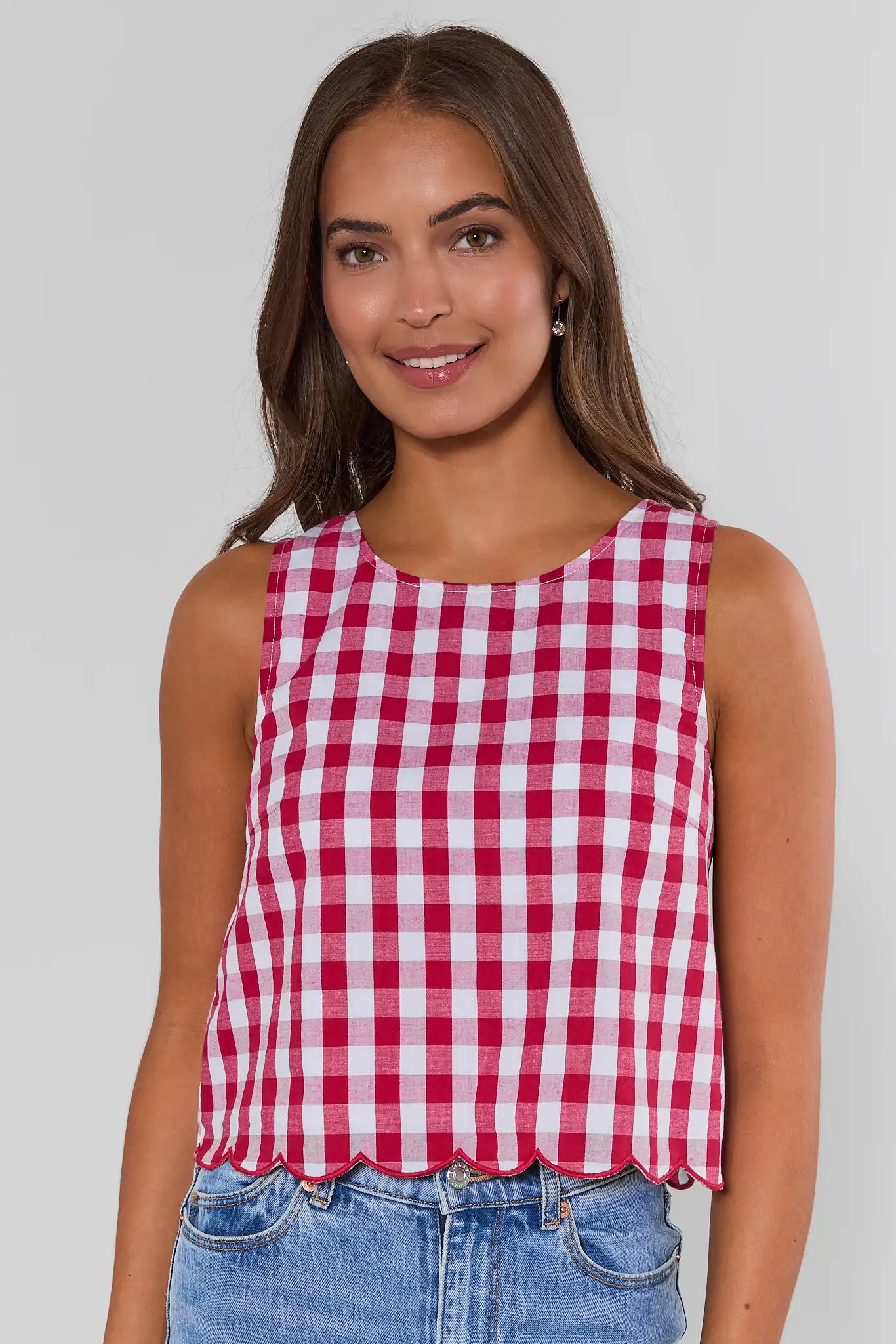 A woman wearing a red and white gingham check sleeveless crop top with a scalloped hem, paired with blue jeans, against a light gray studio background.