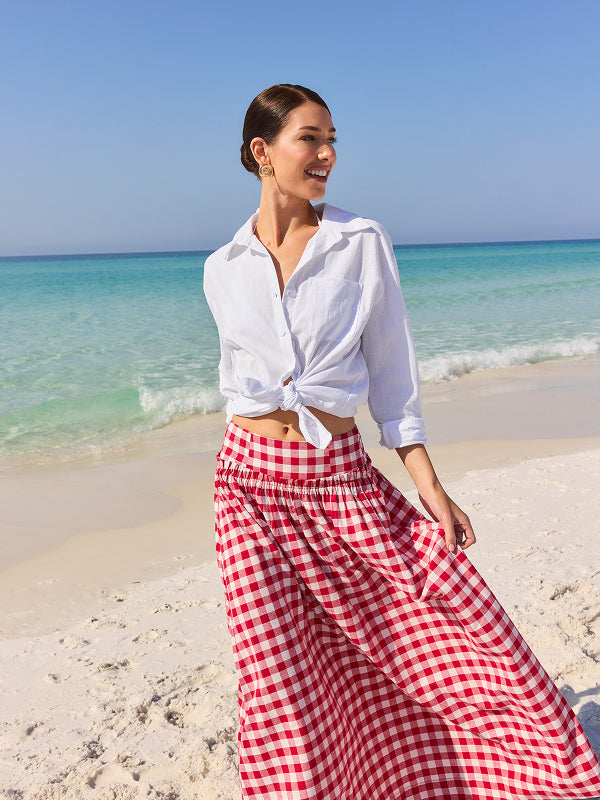 Woman in white shirt and red checkered pants standing on a beach with clear blue water and sky. exper