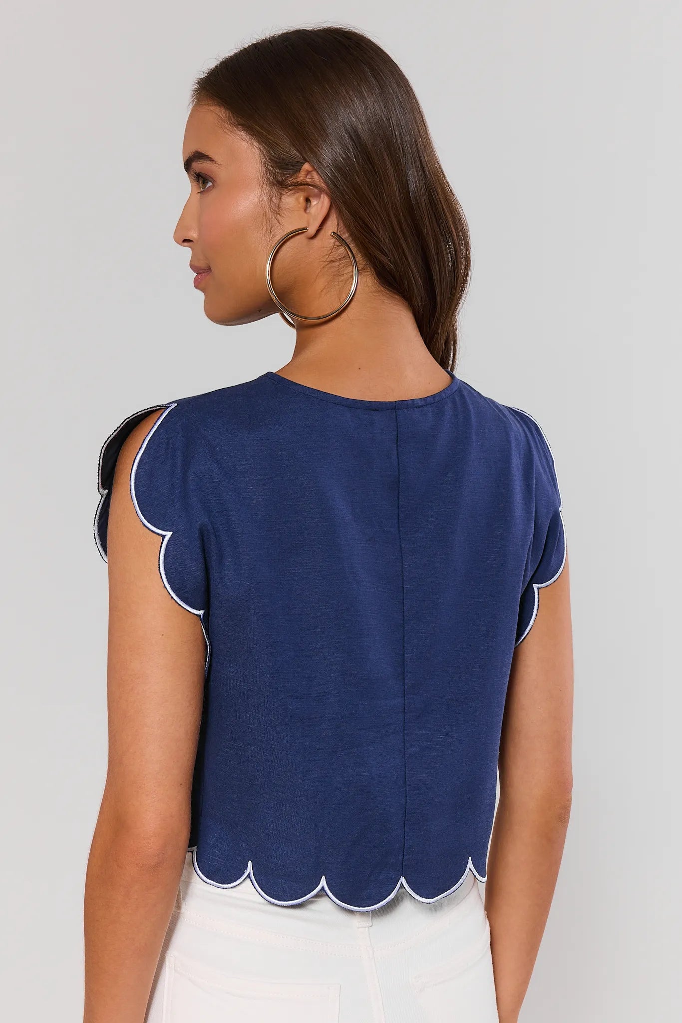 Woman standing wearing a blue top with scalloped edges on a plain background