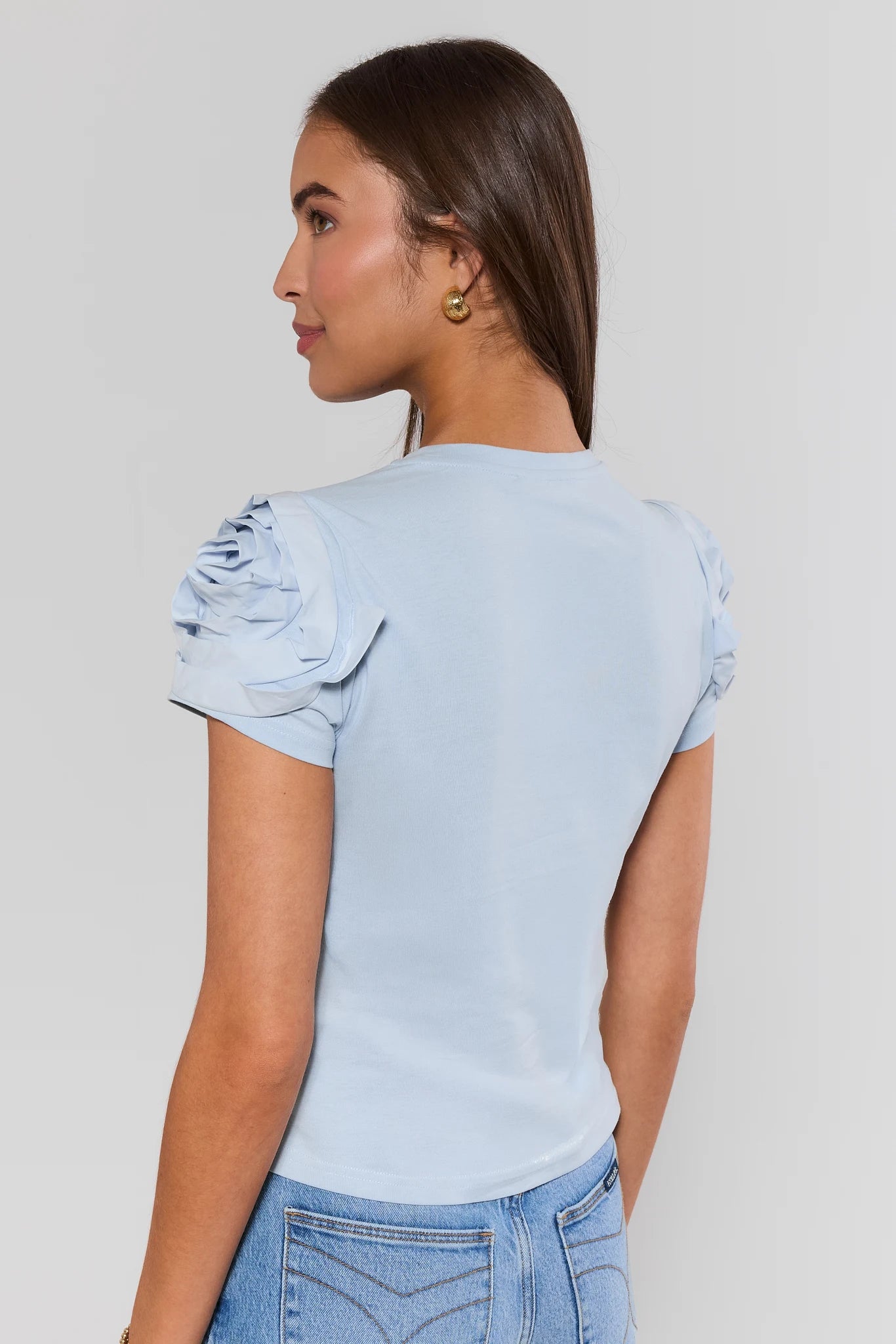 Woman standing wearing a light blue top with ruffled sleeves against a plain background
