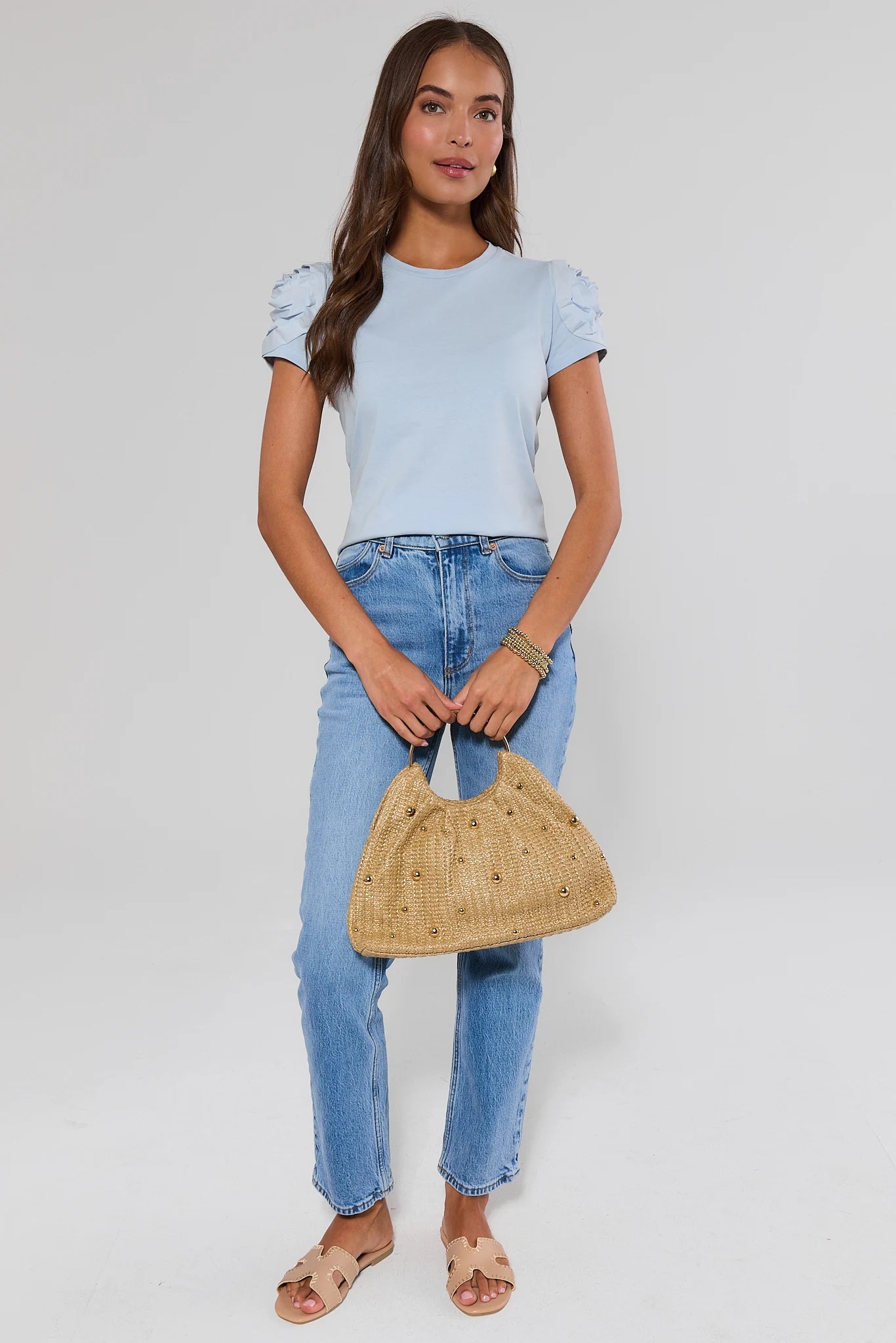Woman wearing a light blue top and jeans, holding a woven handbag on a plain background