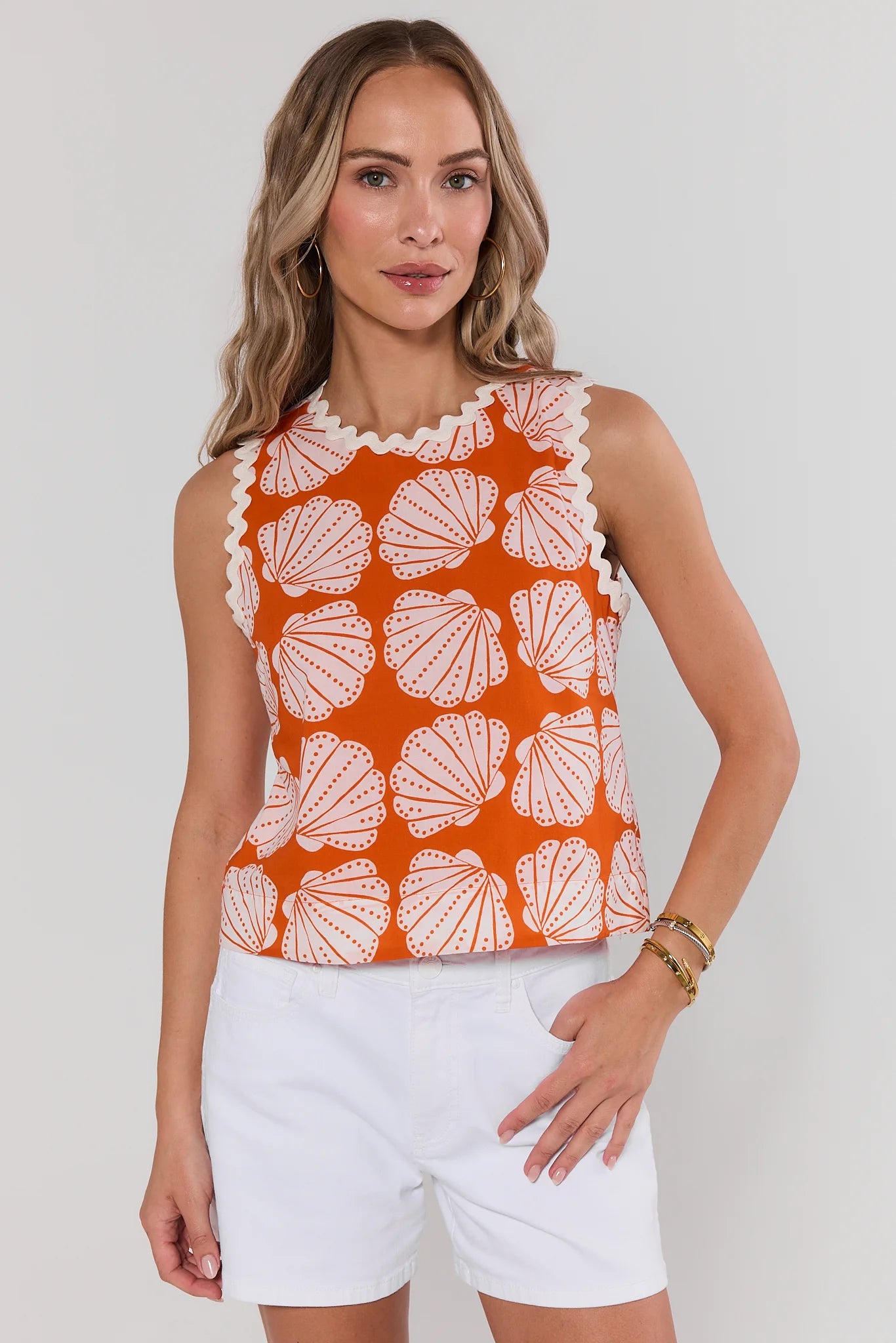 Woman wearing an orange sleeveless top with white shell pattern and white shorts on a plain background