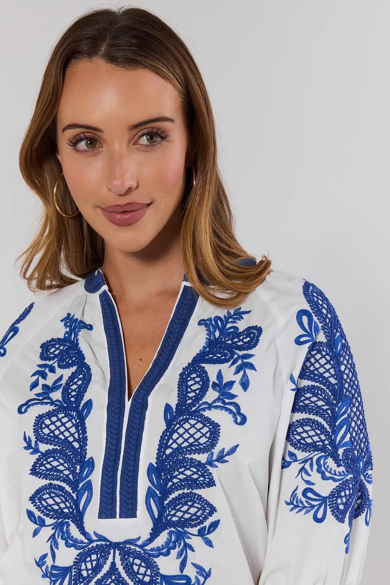 close up Woman wearing a white top with blue embroidery against a plain background