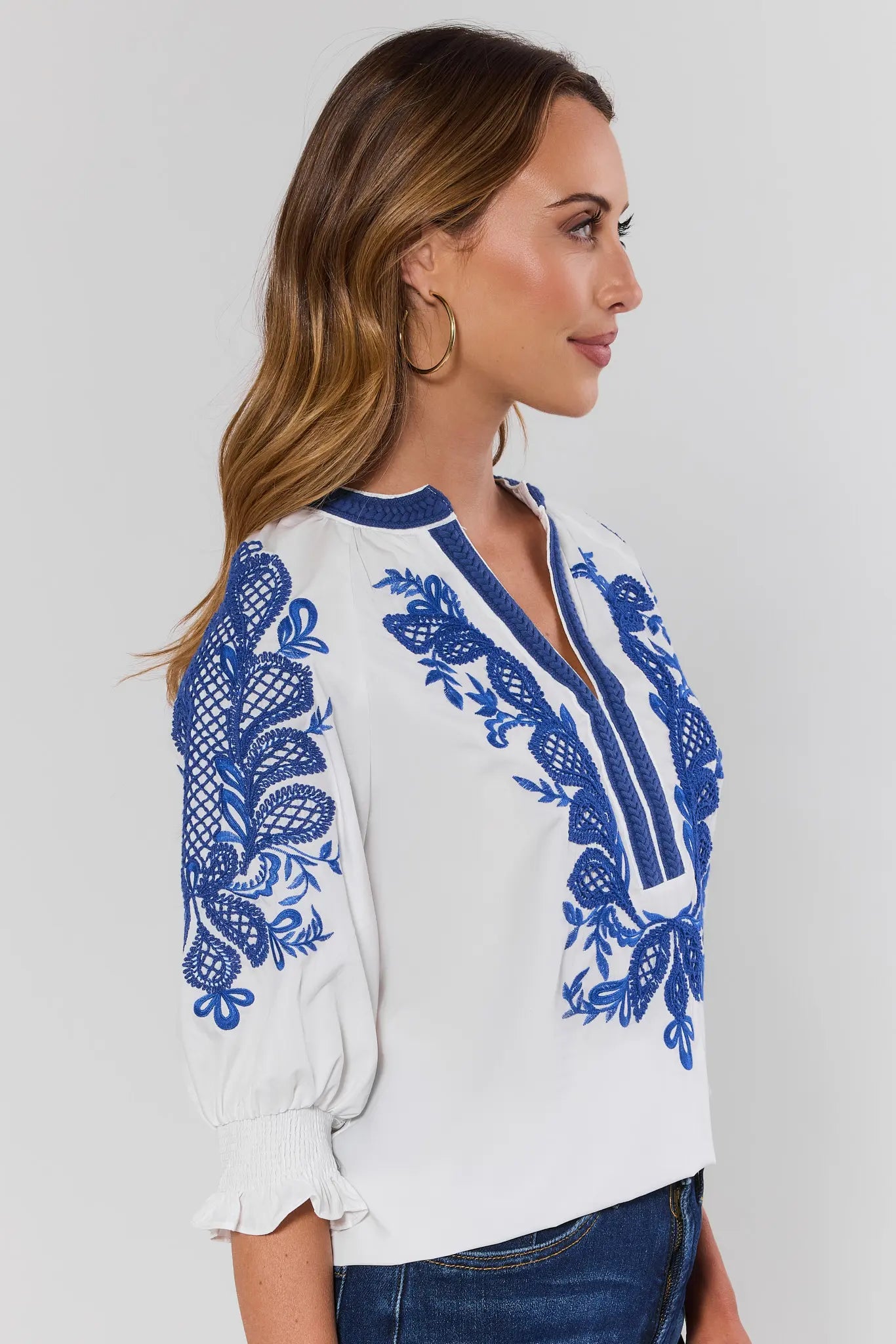 side shot Woman wearing a white blouse with blue embroidery on a plain background