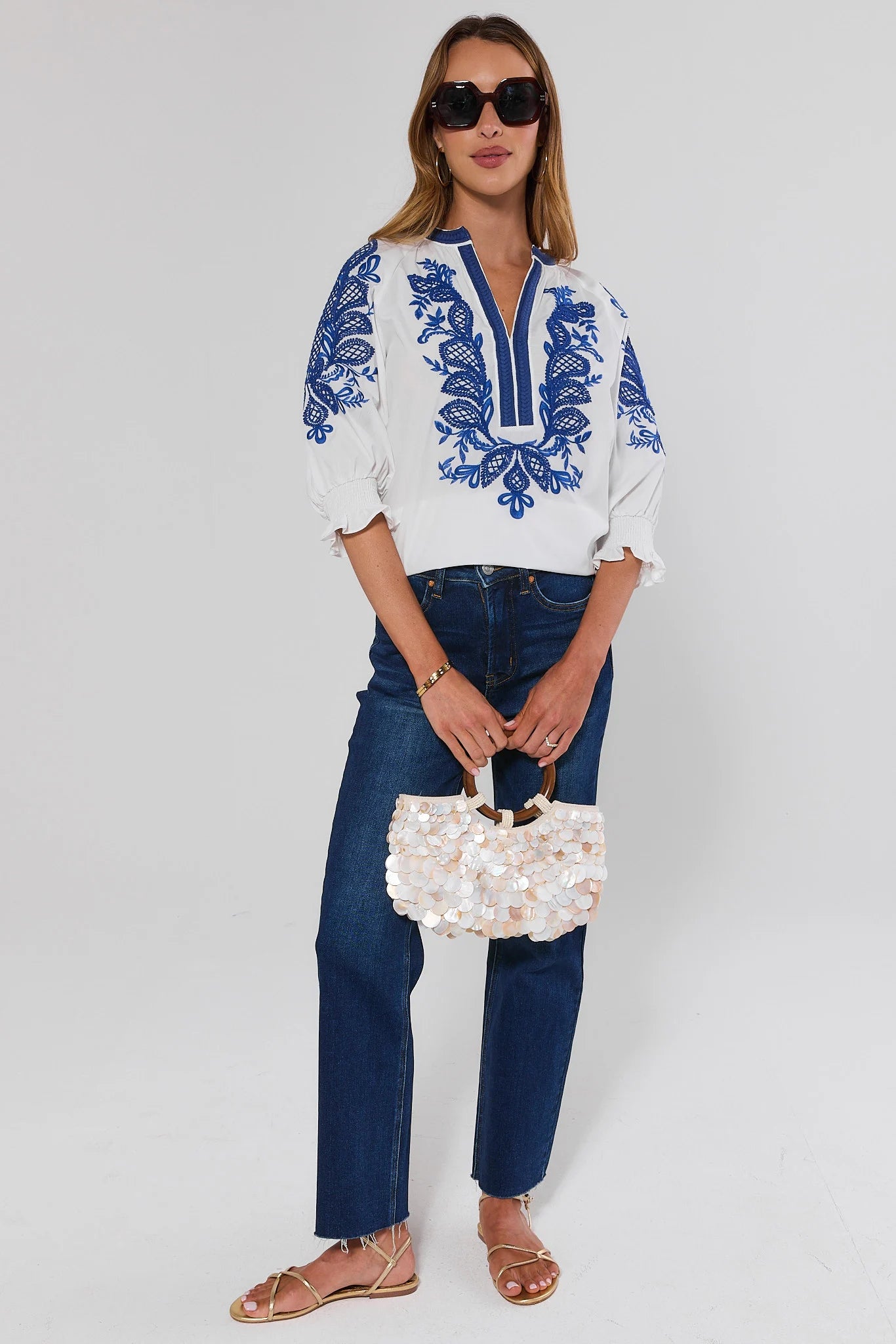 Woman wearing a white blouse with blue embroidery and jeans, holding a white handbag and wearing sunglasses