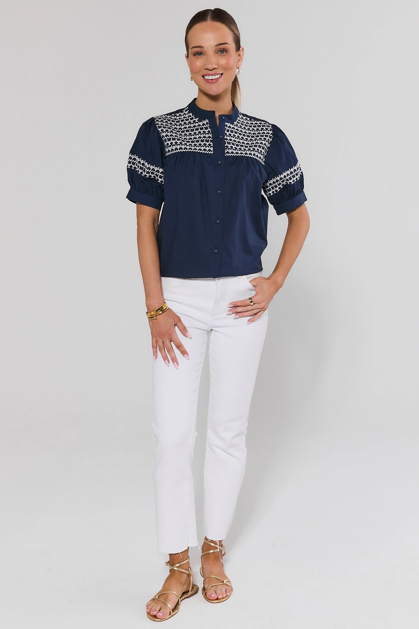 Woman standing wearing a navy blouse with white patterns and white pants on a light gray background