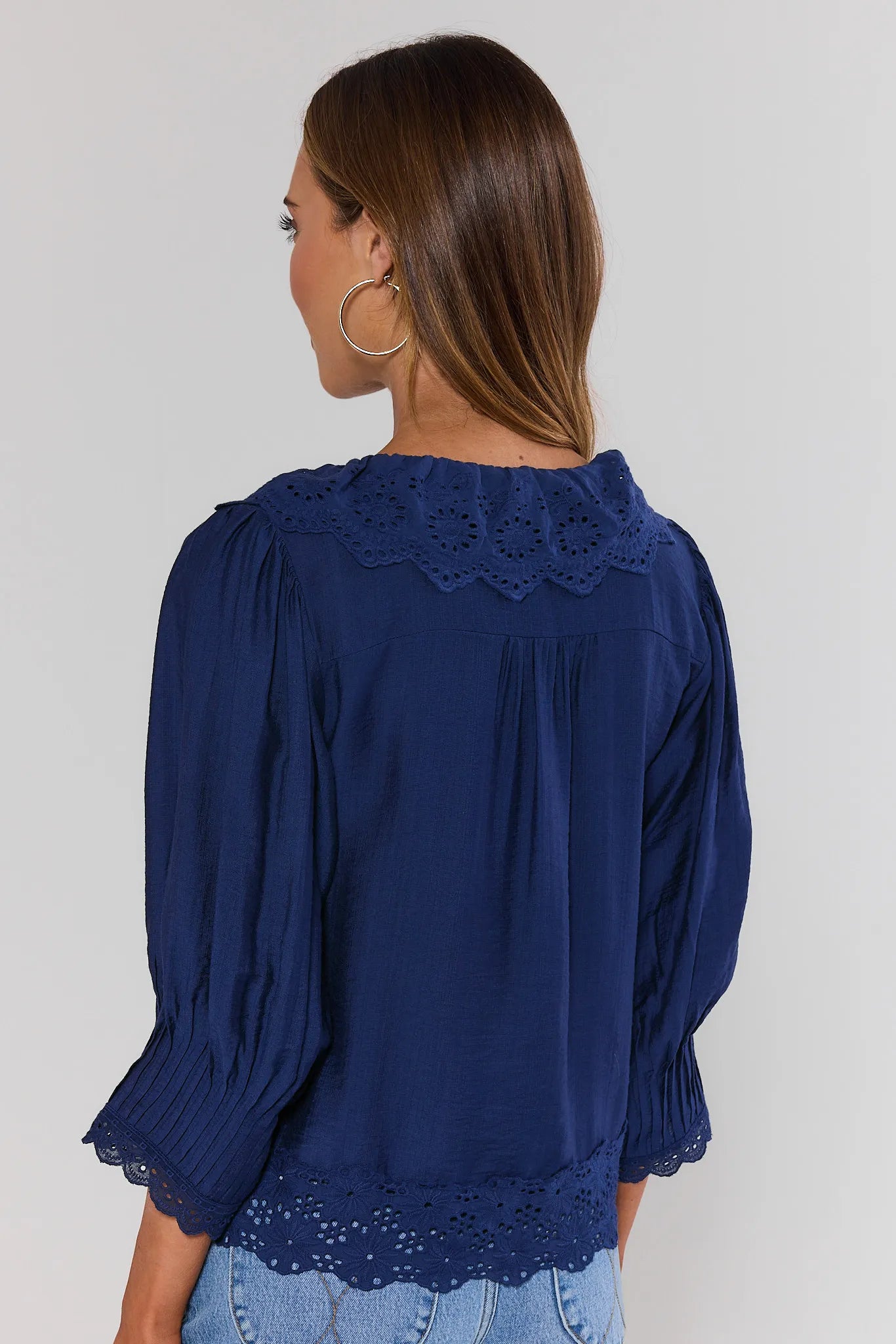 Woman standing wearing a blue blouse with lace details on a plain background