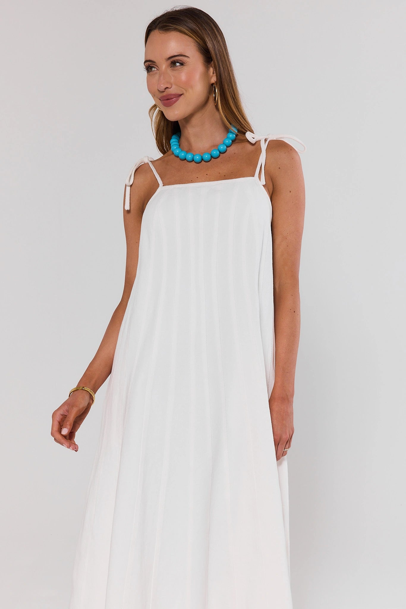 Woman wearing a white dress with thin straps and a blue necklace on a plain background