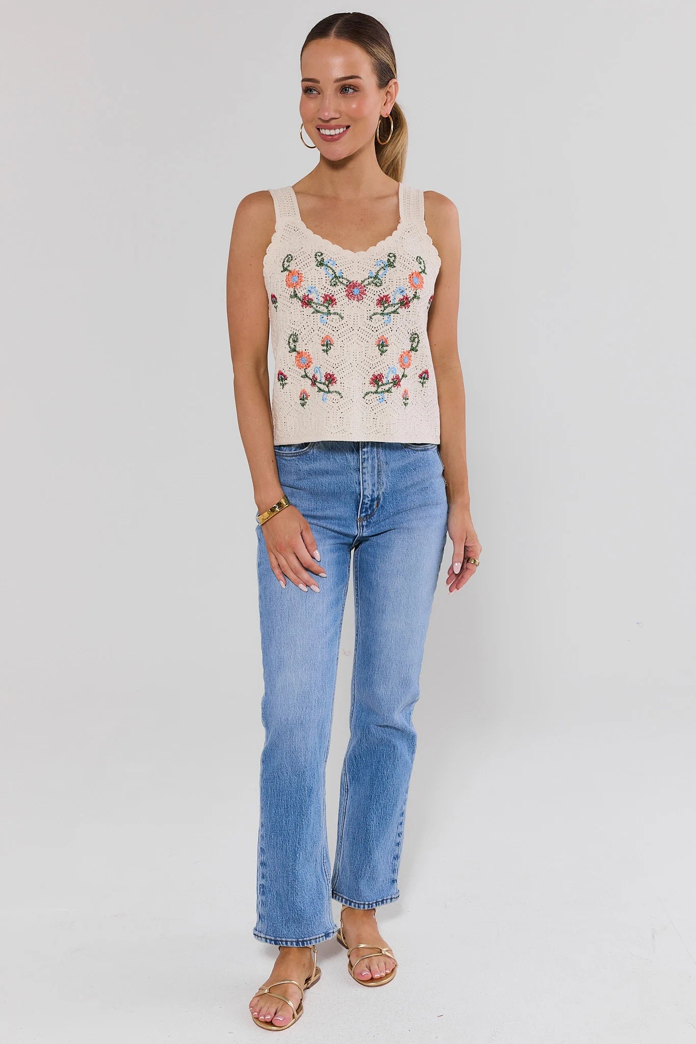Woman standing wearing a floral embroidered tank top and blue jeans on a white background