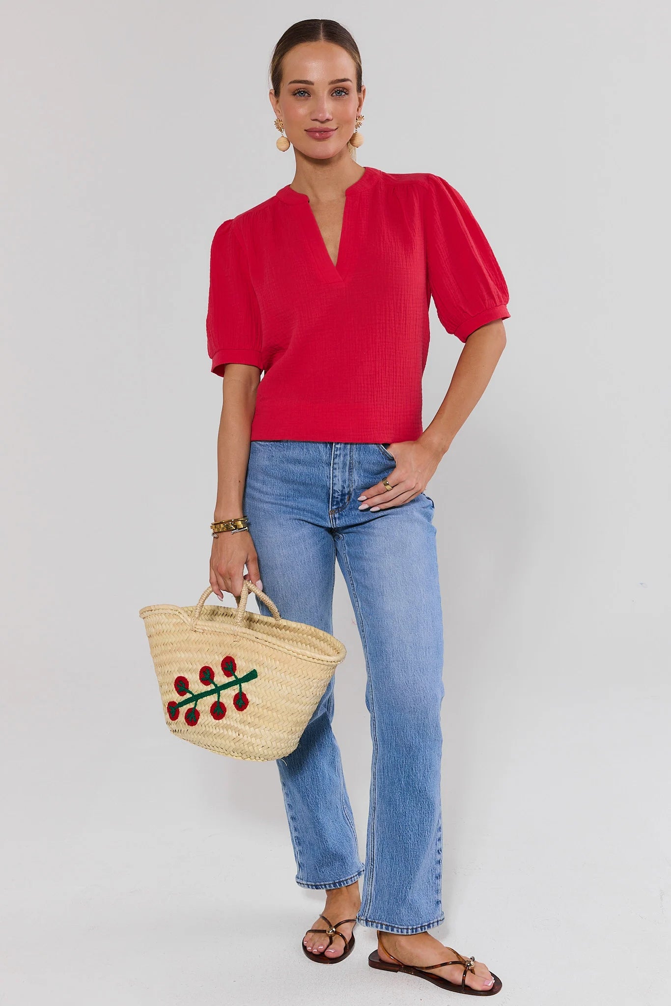 Woman wearing a red blouse and blue jeans, holding a straw bag with floral details, on a plain background.