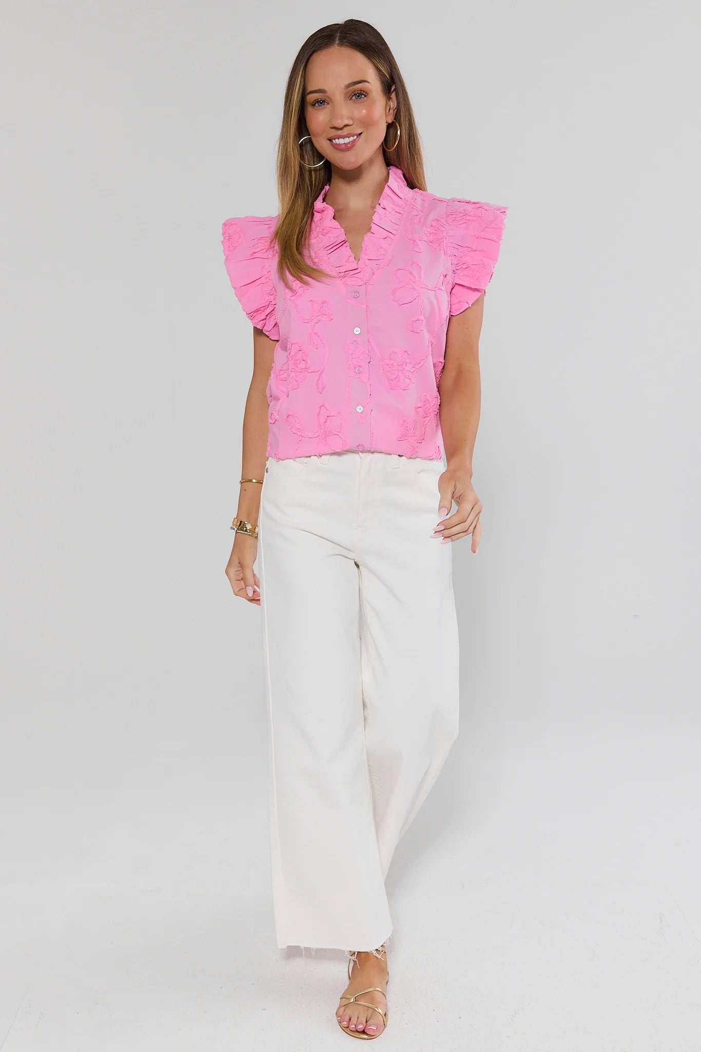 Woman wearing a pink blouse with ruffled sleeves and white pants on a gray background stand