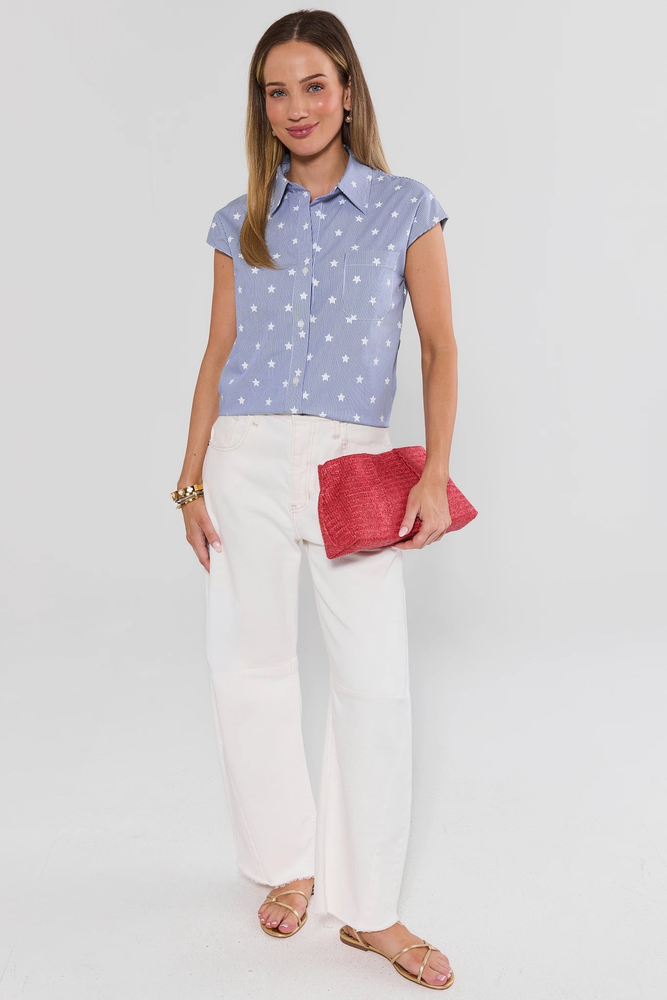 Woman wearing a blue patterned shirt, white pants, and holding a red clutch against a plain background