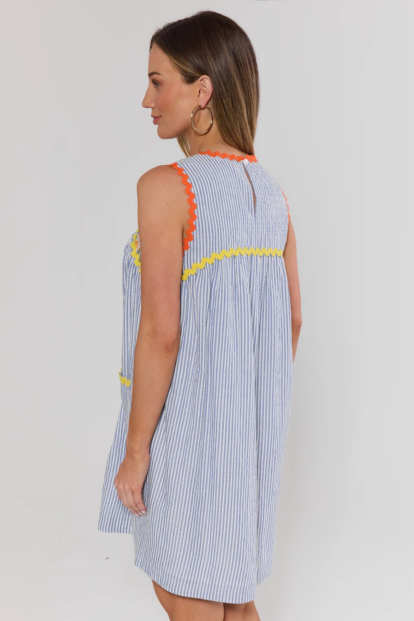 Woman standing wearing a sleeveless dress with striped pattern and colorful trim on a plain background 