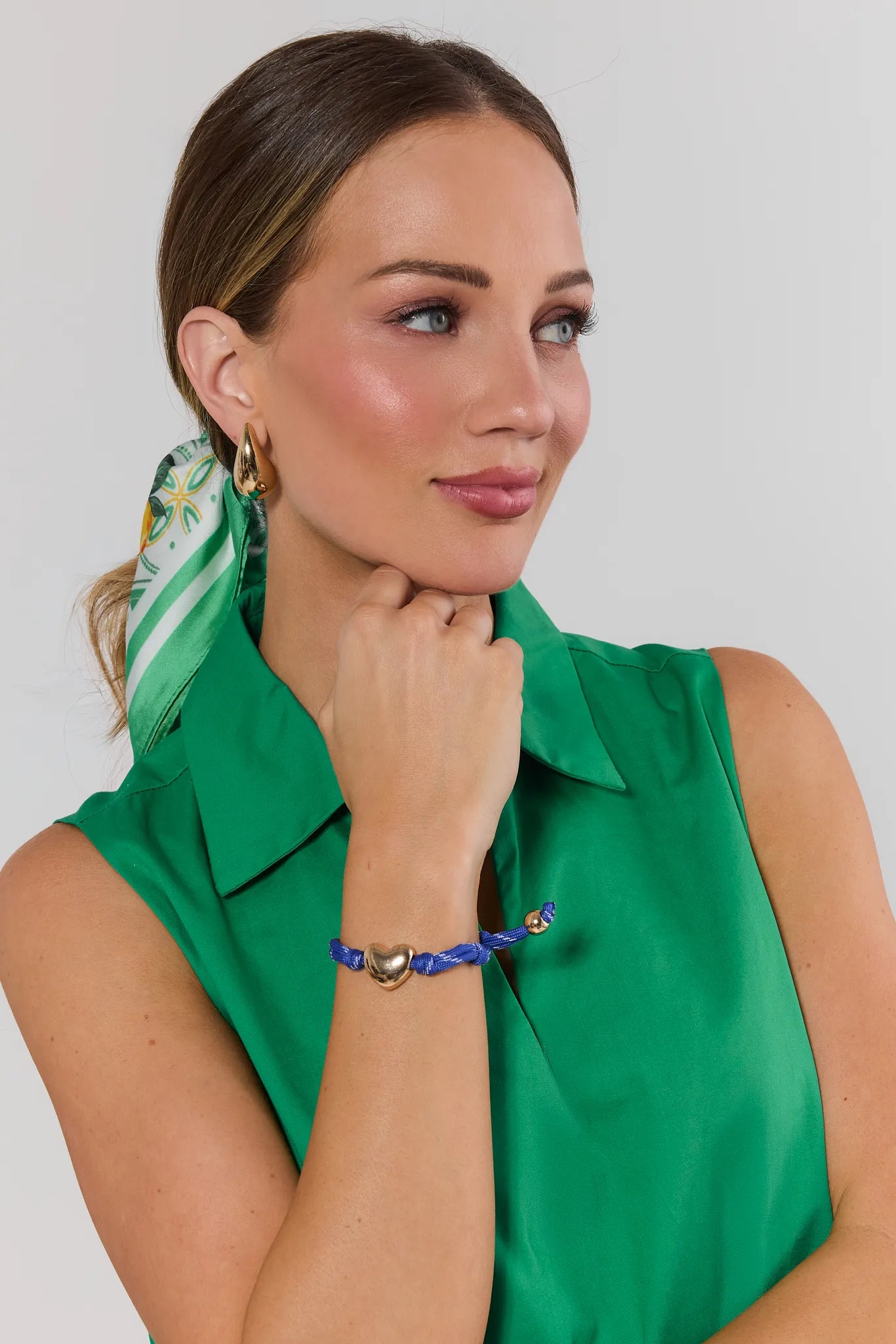 Woman wearing a green sleeveless top with a green scarf and blue bracelet against a white background