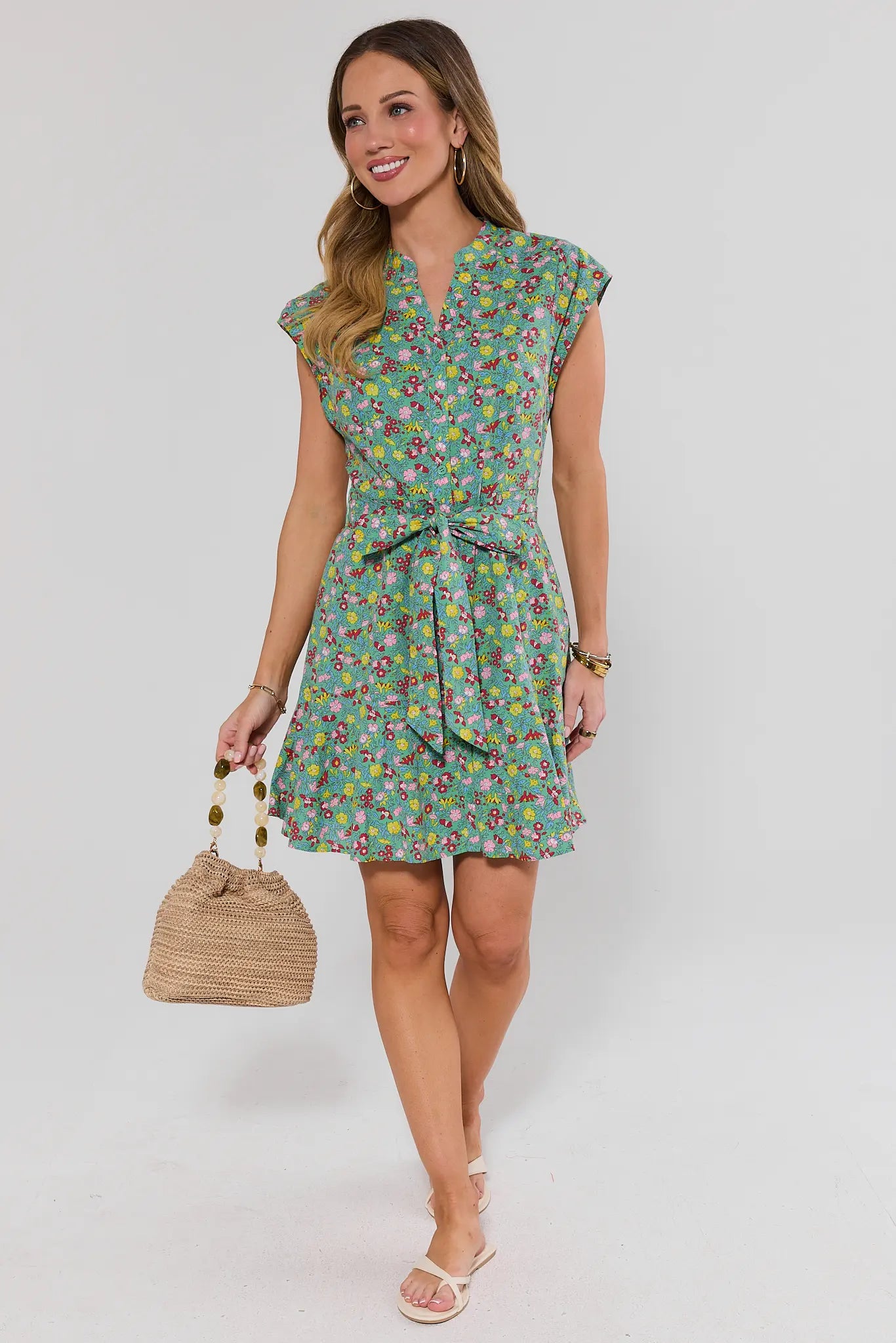 Woman standing wearing a floral dress holding a woven bag on a plain background