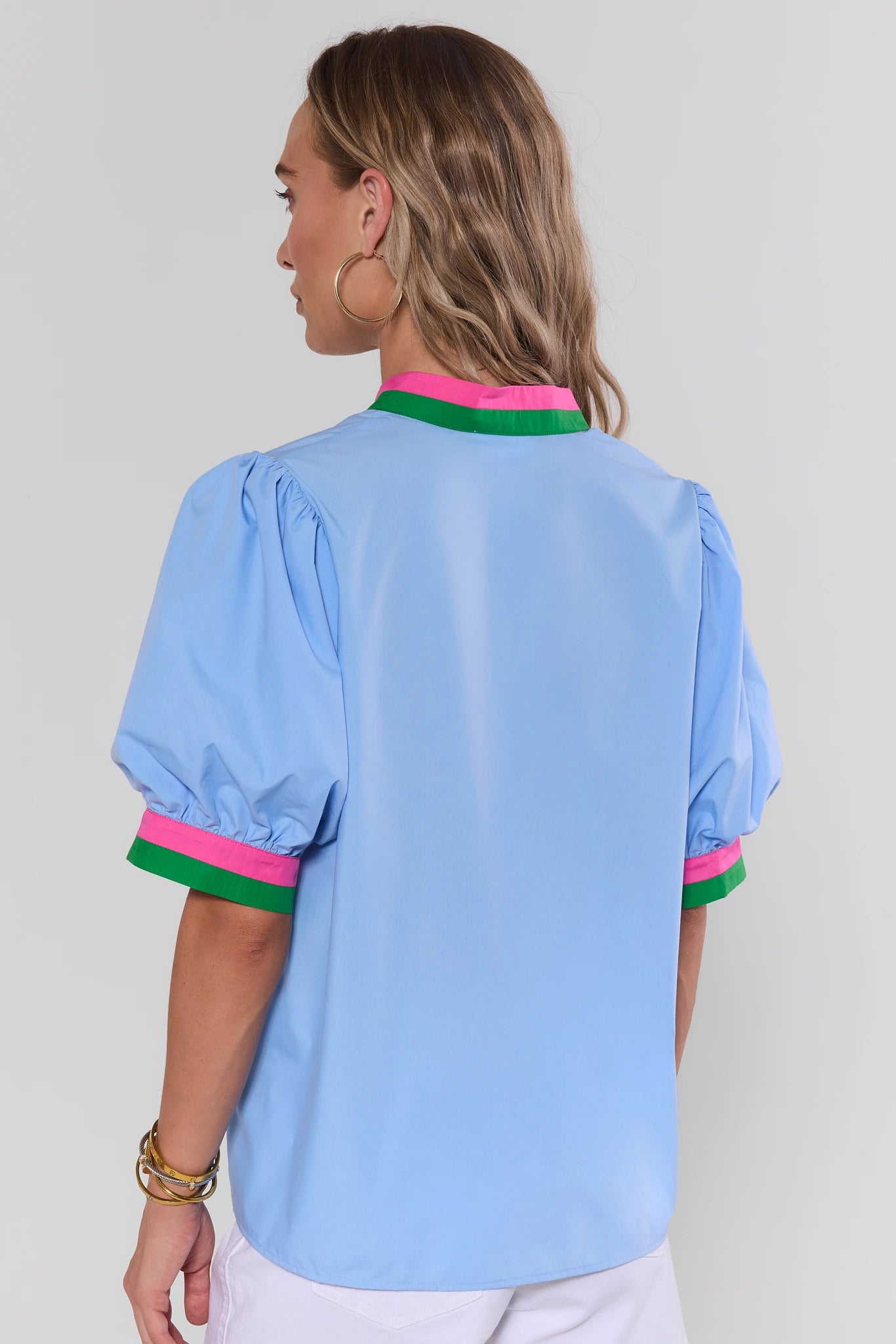Woman standing wearing a light blue blouse with pink and green trim on a plain background