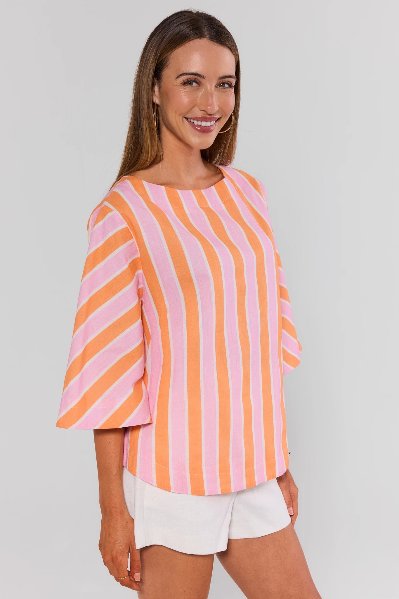 Woman wearing a pink and orange striped top with white shorts on a plain background side