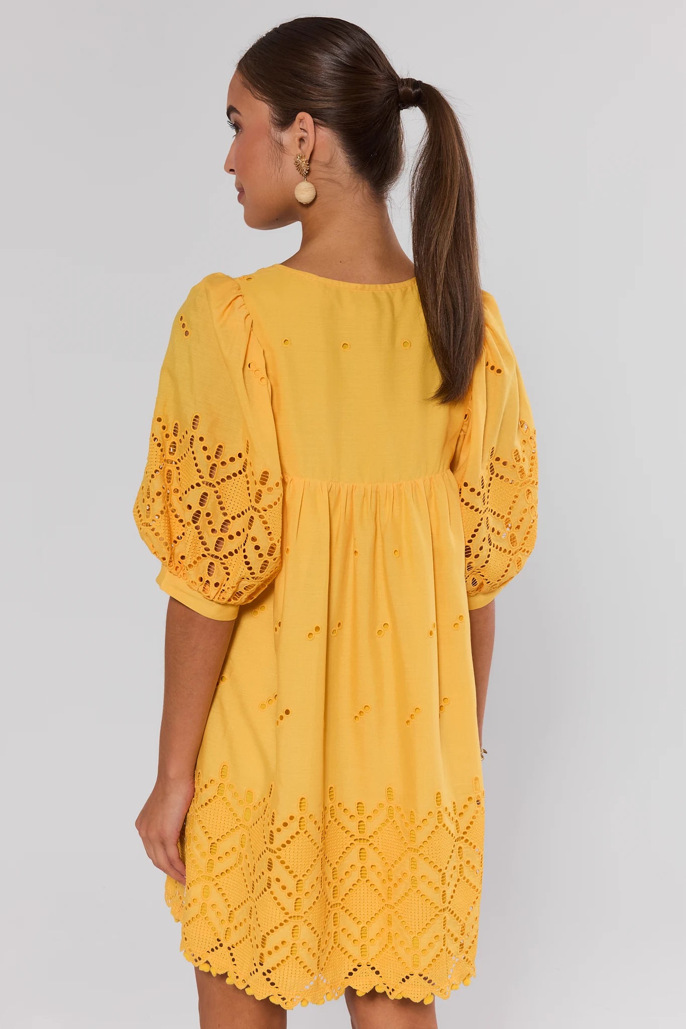 Woman wearing a yellow dress with lace details on a plain background stand