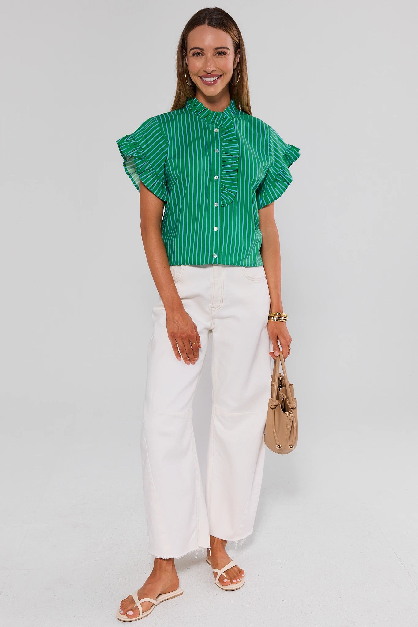 Woman wearing a green striped shirt and white pants, holding a beige handbag on a light gray background