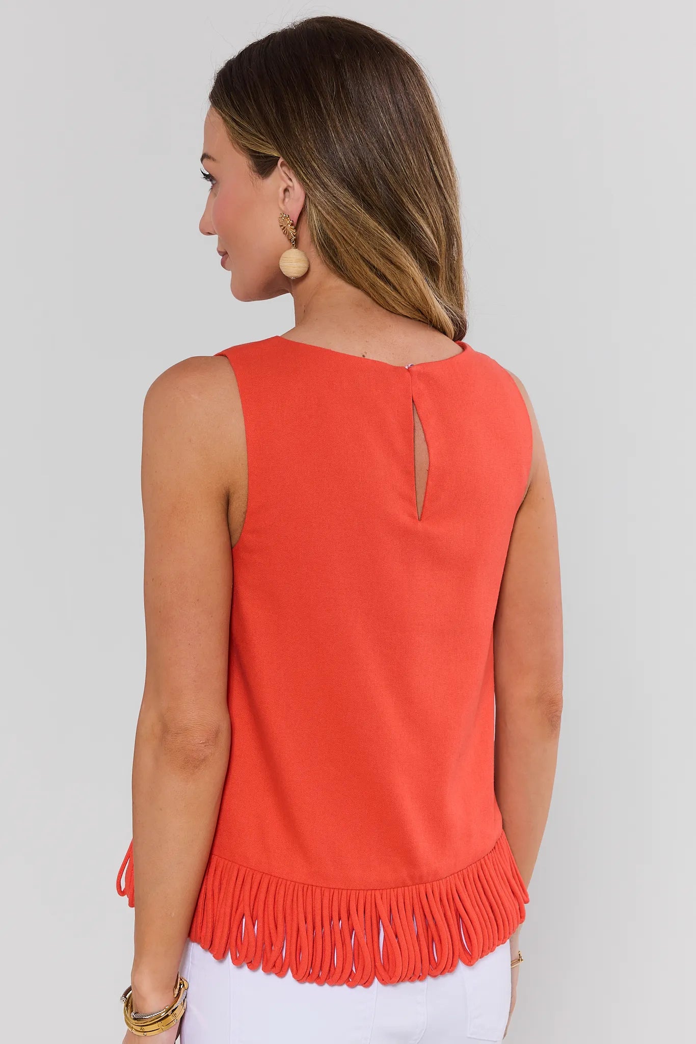 Woman standing wearing an orange sleeveless top with fringe details on a plain background