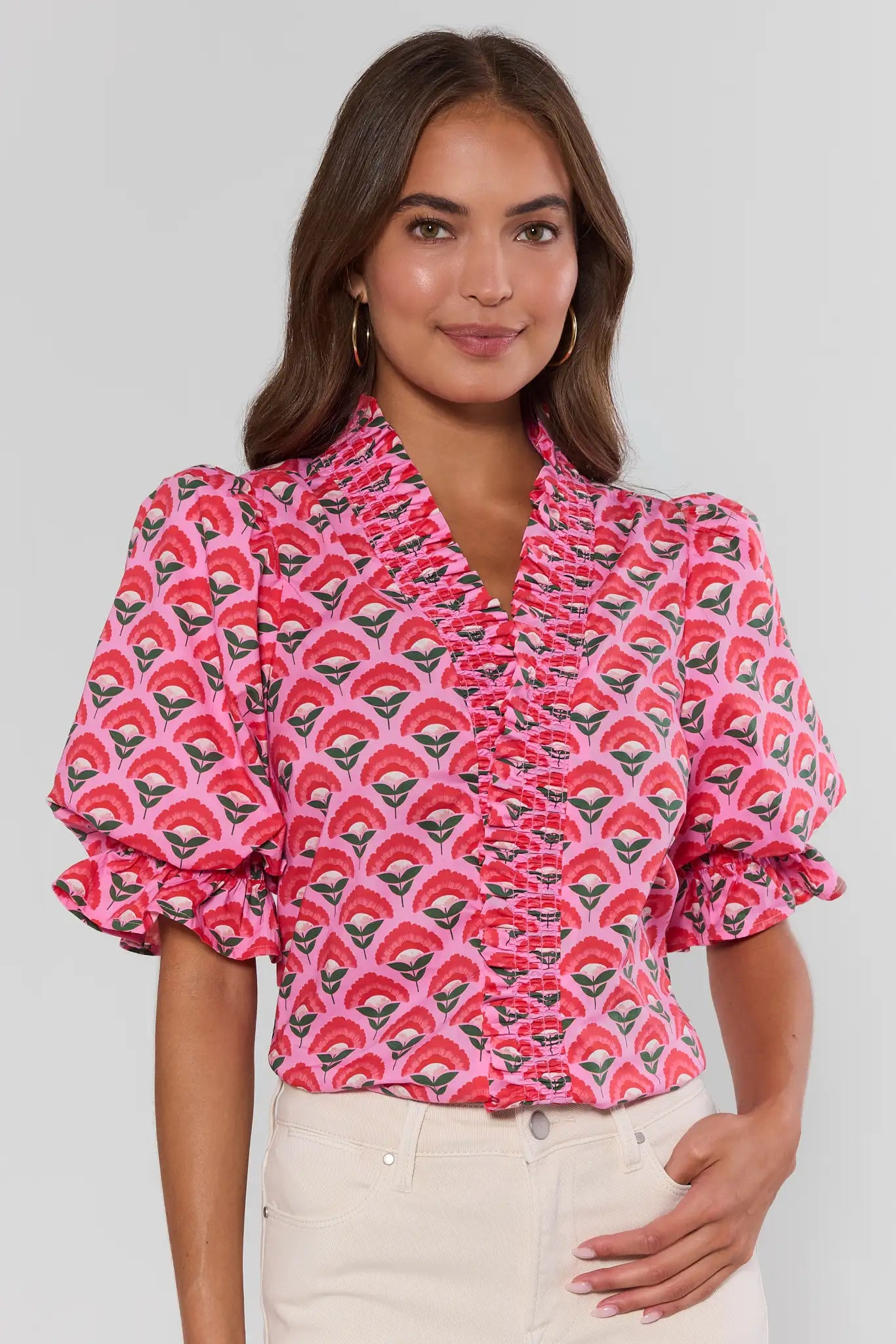 Woman wearing a pink patterned blouse with white pants on a plain background