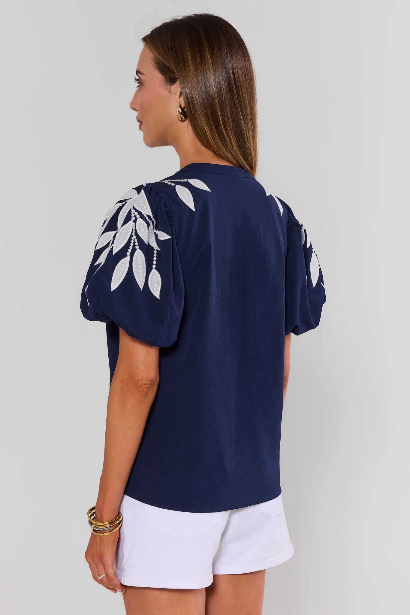 Woman standing wearing a navy blue top with white leaf patterns and white shorts on a light gray background