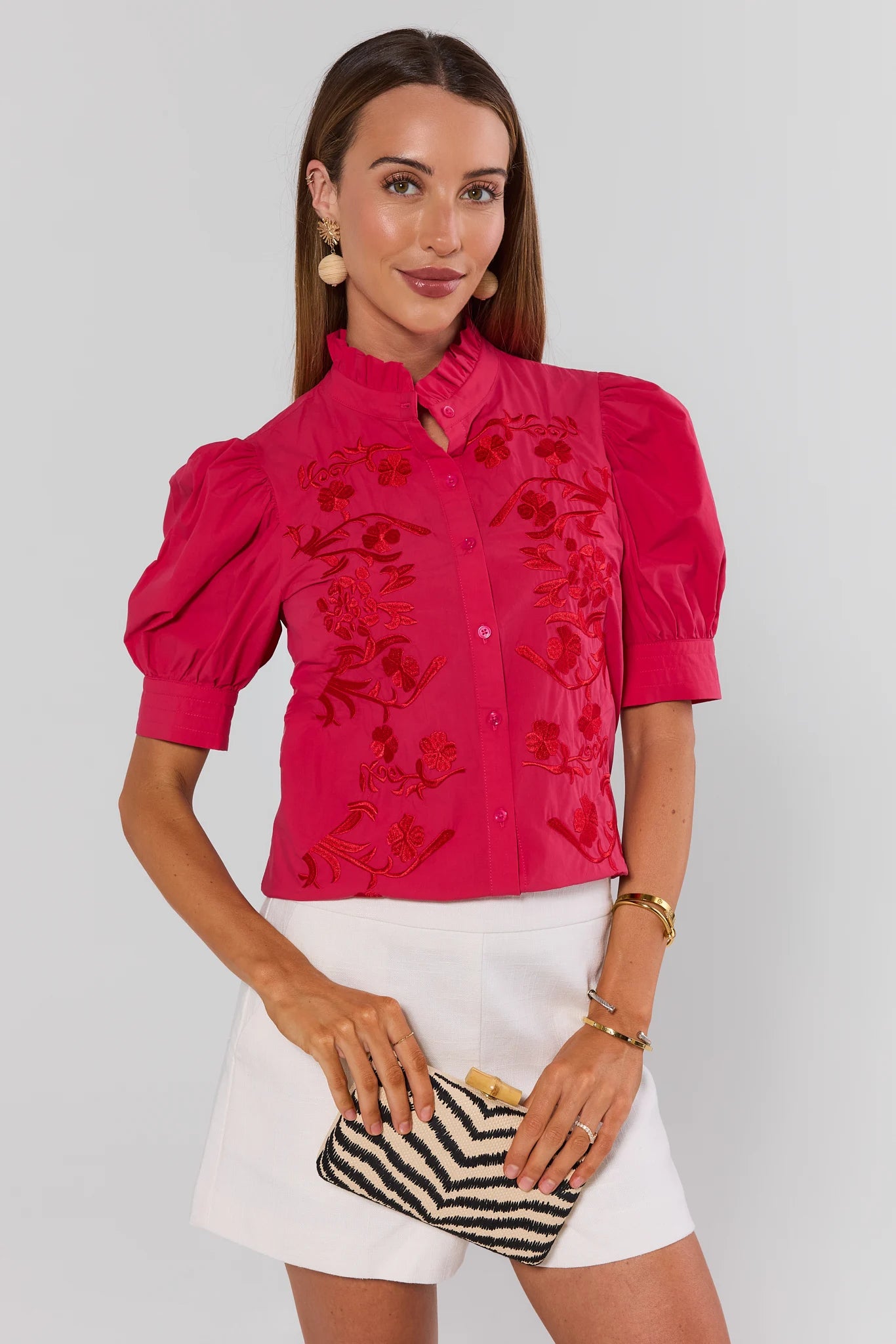 Woman wearing a pink blouse with floral embroidery and white shorts, holding a striped clutch.