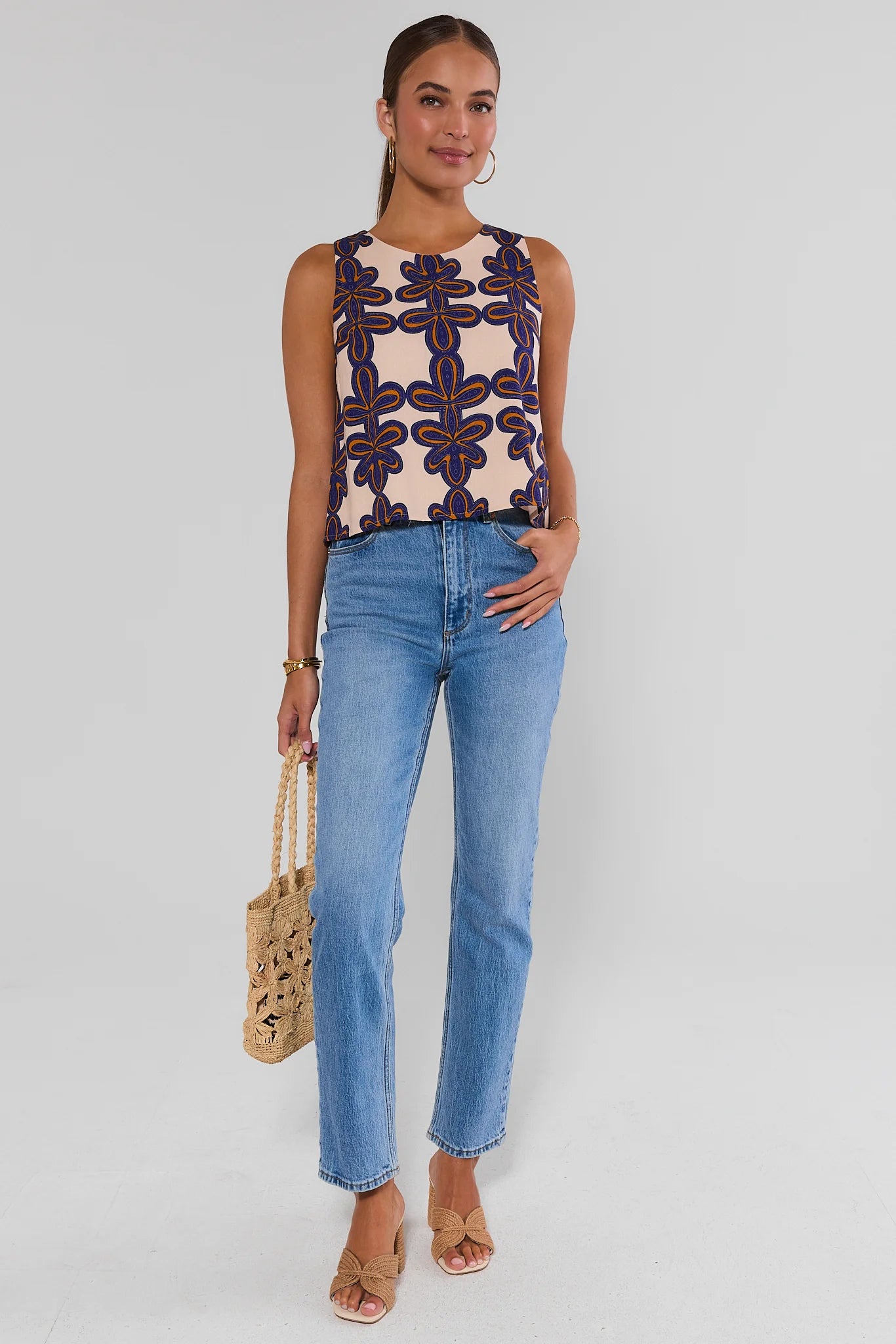 Woman wearing a sleeveless patterned top and blue jeans, holding a straw bag on a white background standing